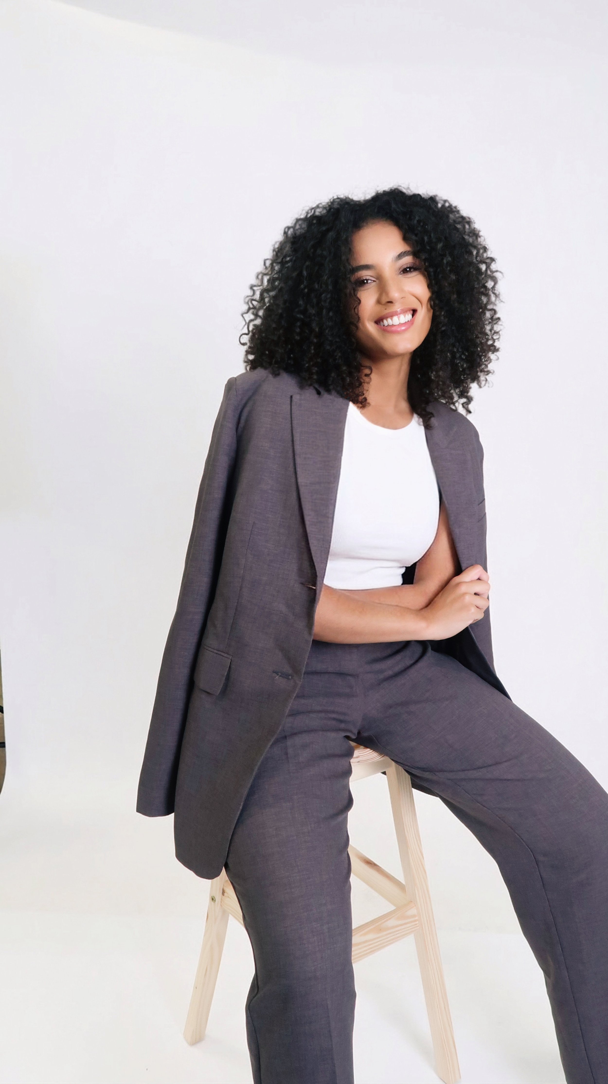 A woman with curly black hair wearing a gray suit and white top, sitting on a light wooden stool against a plain white background, smiling.