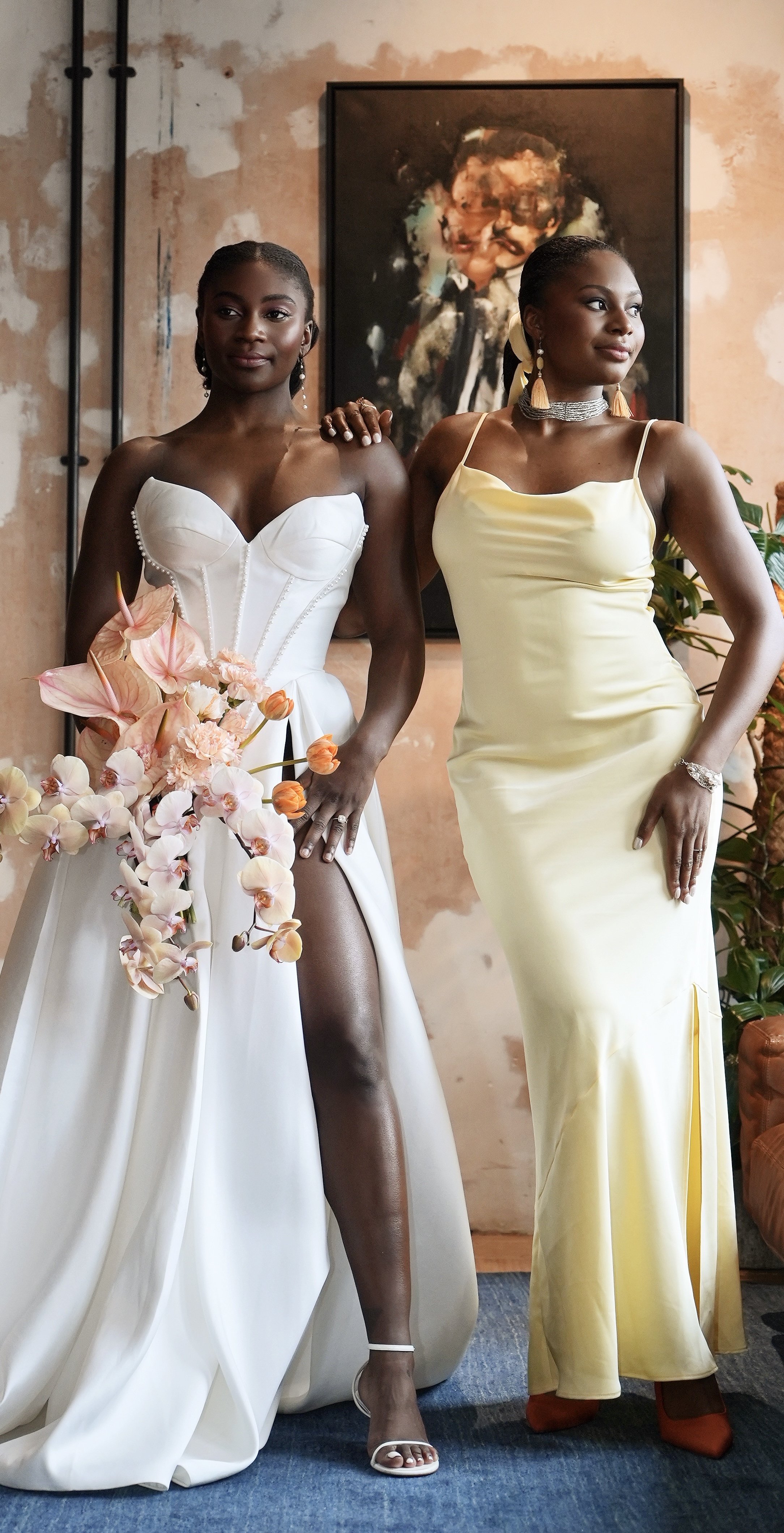 Two women stand side by side; the woman on the left wears a white wedding gown with a slit showing her leg, holding a bouquet of pink and orange flowers, and the woman on the right wears a yellow dress with jewelry, in a room with an abstract painting and plants.