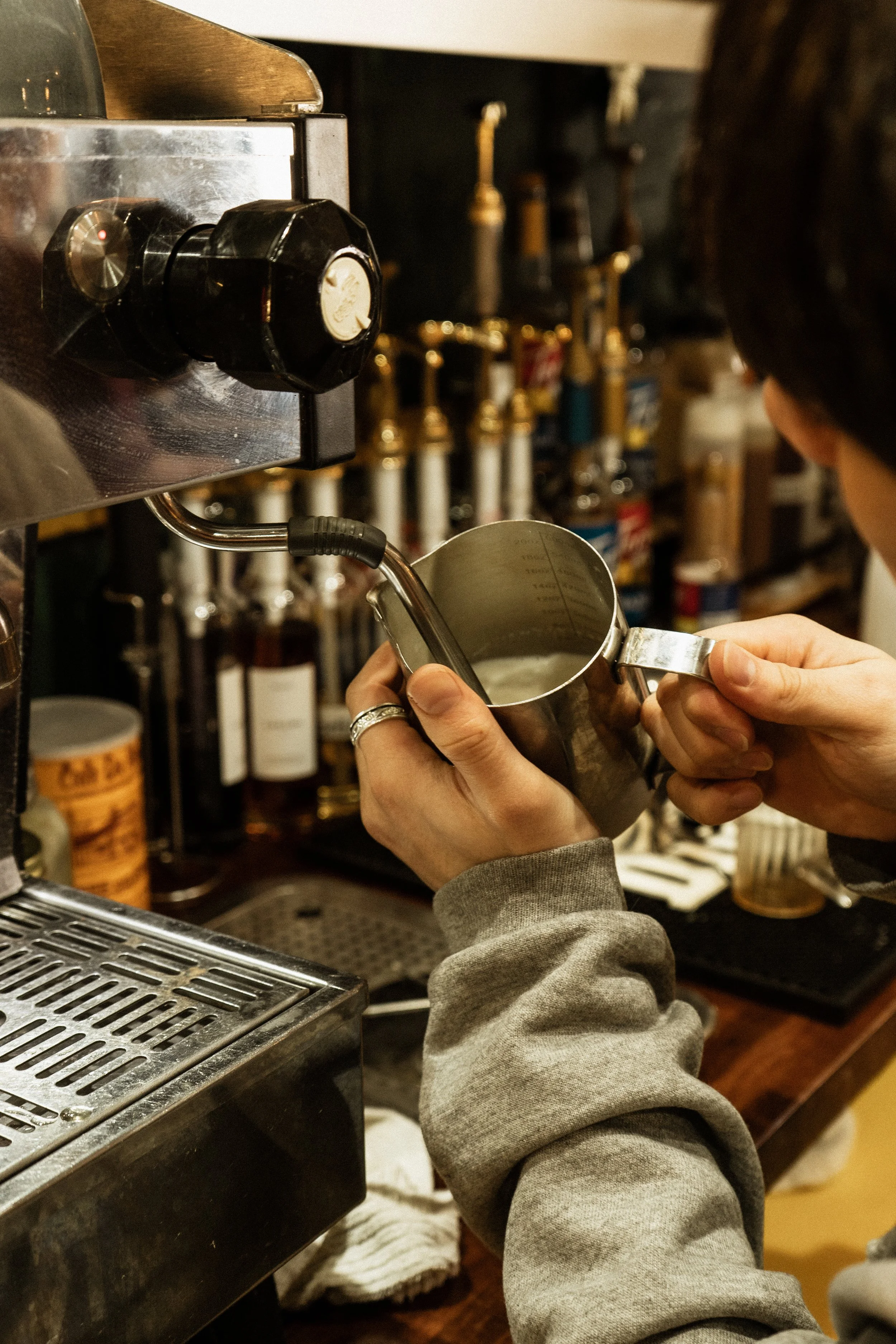 A person holding a metal pitcher under a coffee machine to collect coffee or espresso, with various bottles and condiments in the background.