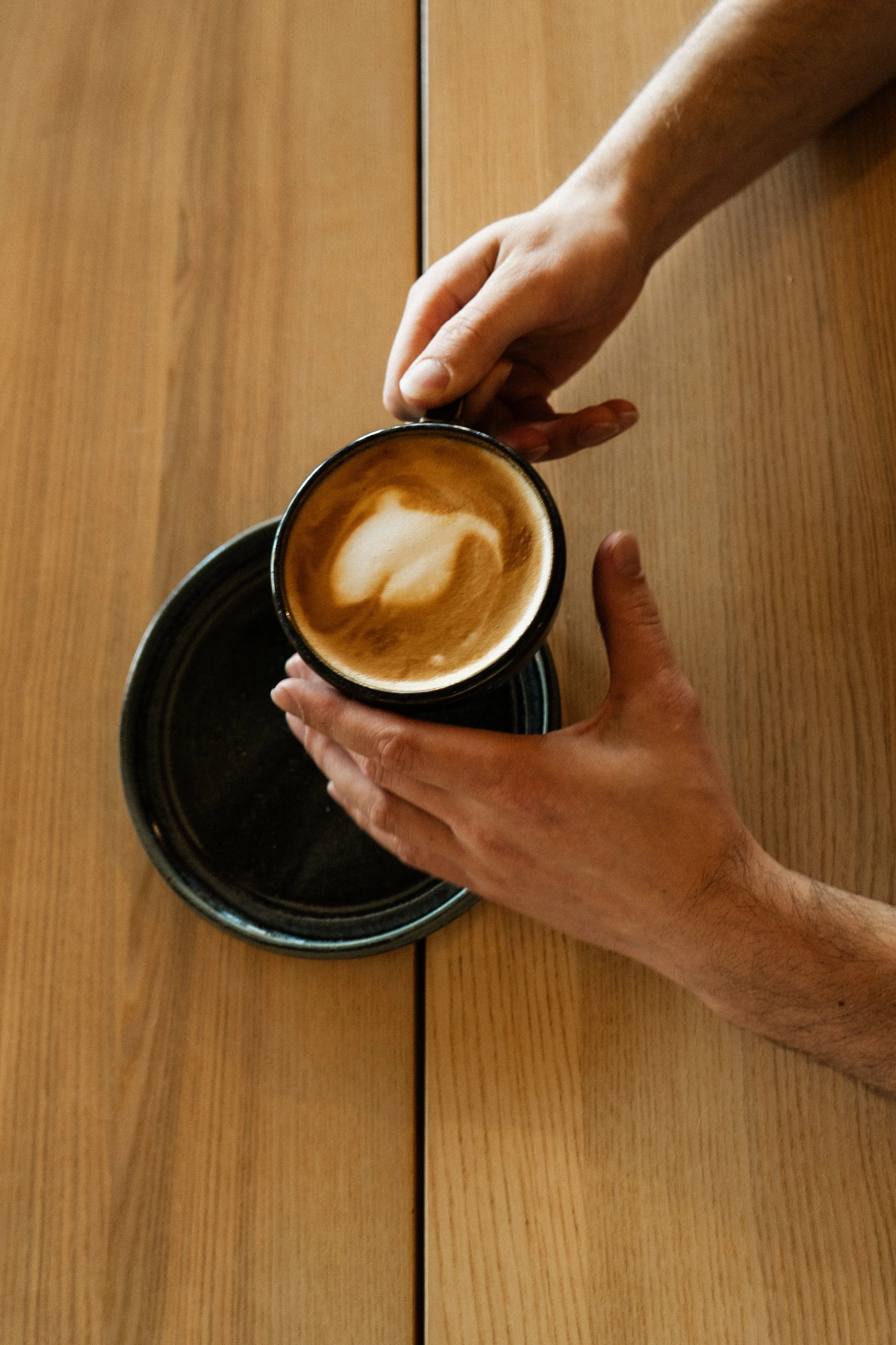 A person holding a coffee cup with latte art, sitting at a wooden table with another empty cup beneath.