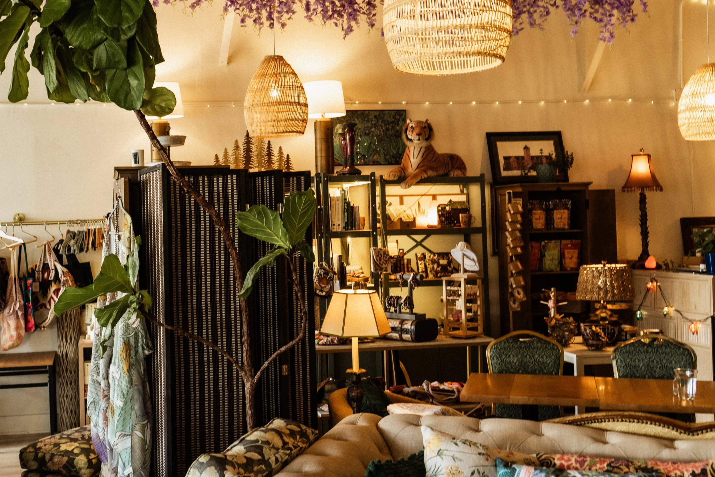 Interior of a cozy store or cafe with hanging wicker lights, a plush beige sofa, plants, and decorative items, including a stuffed tiger on a shelf, framed pictures, and various goods displayed on shelves.