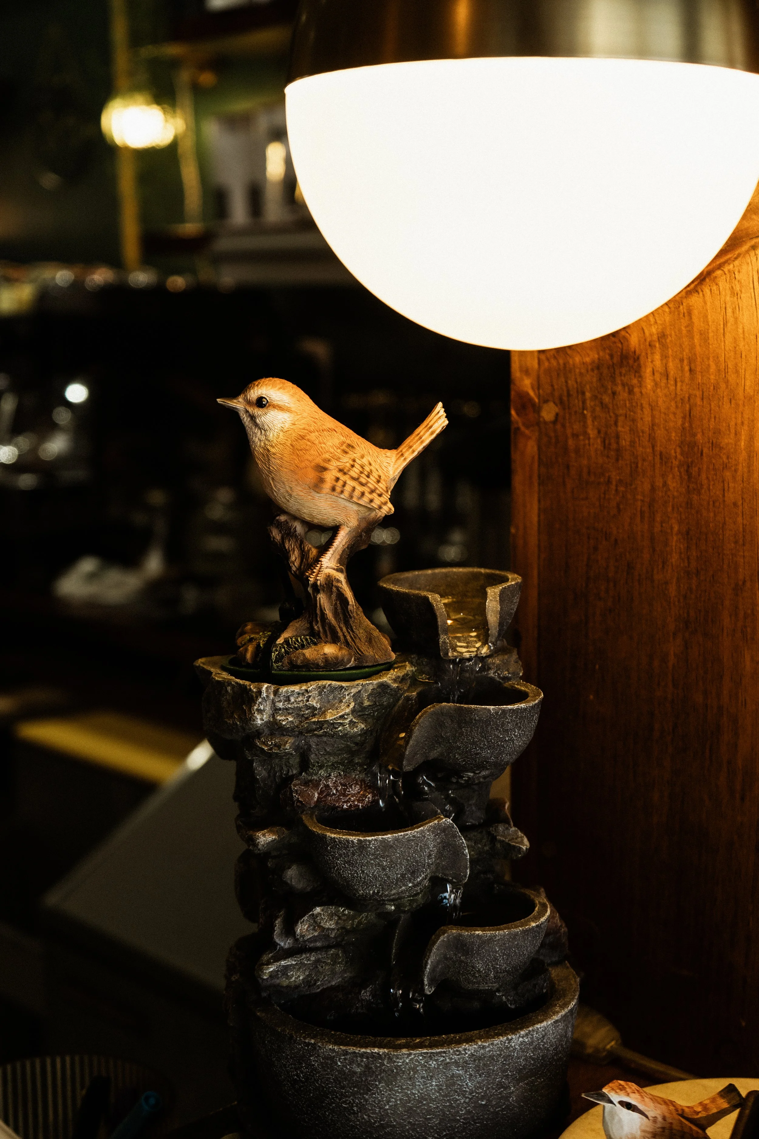 A small decorative bird sculpture perched on a branch, next to a multi-tiered black stone fountain with water, illuminated by a bright white wall-mounted light fixture in a dimly lit room.