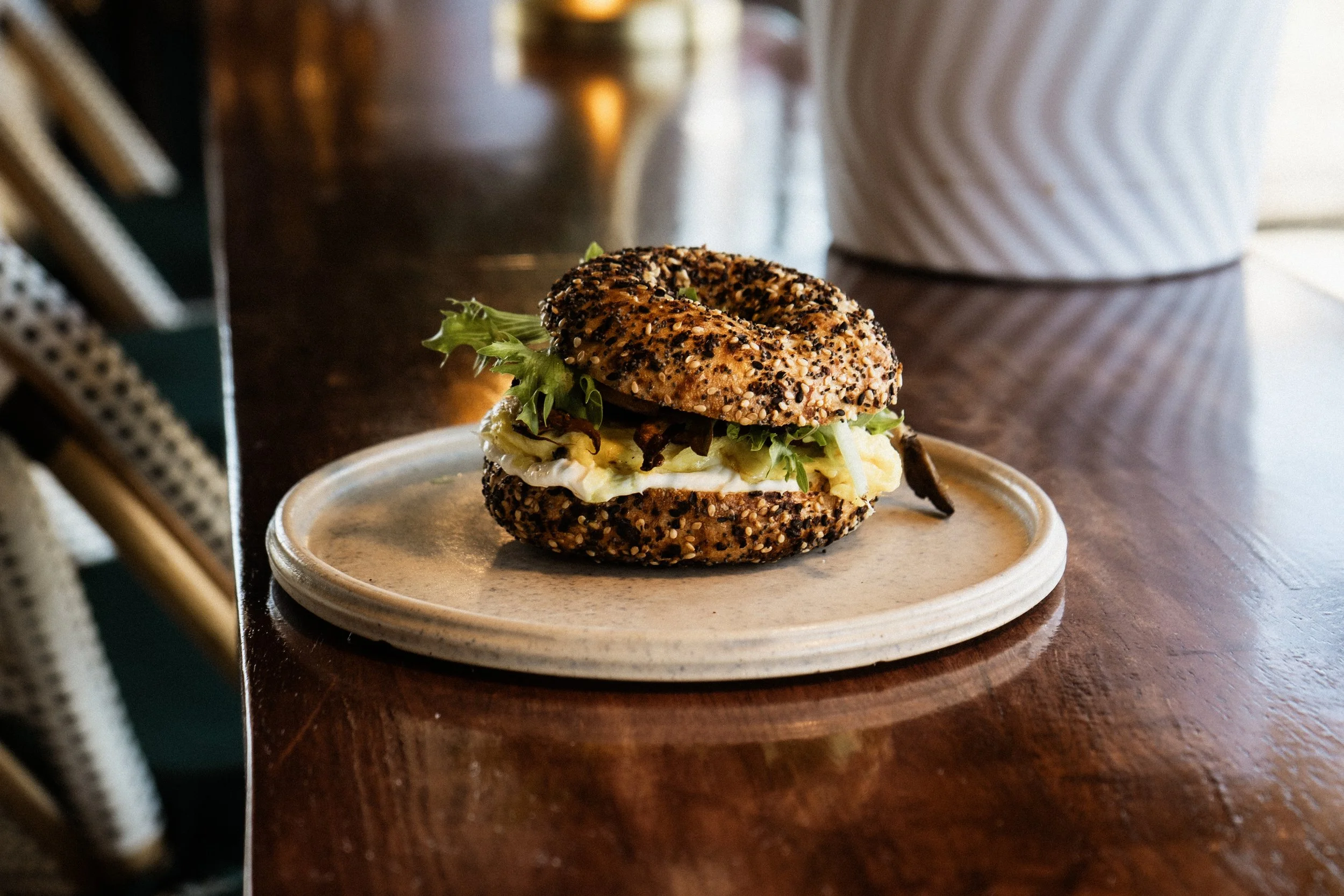 A bagel sandwich on a ceramic plate with lettuce, egg, and meat, topped with a sesame and poppy seed bagel, resting on a dark wooden table.