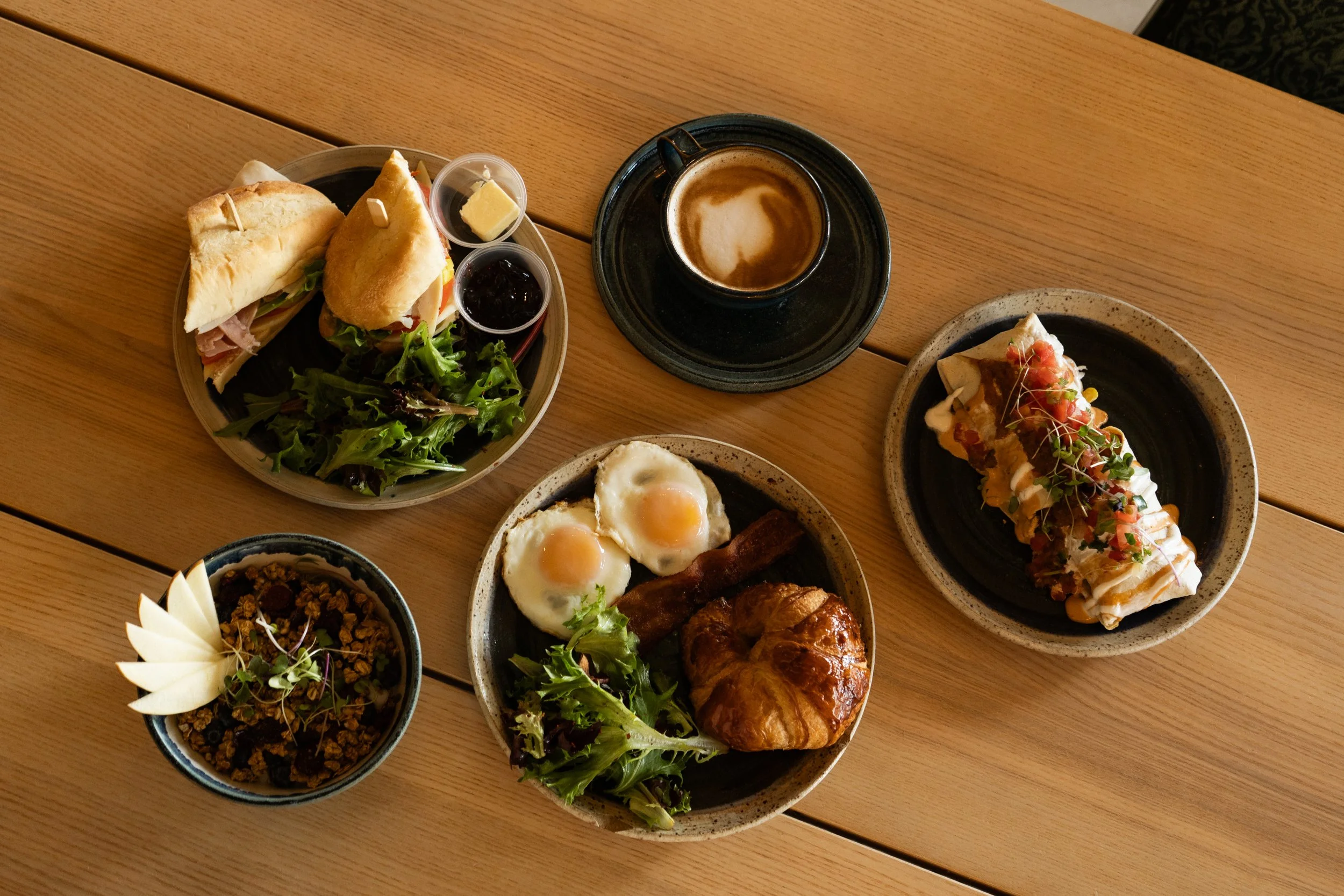 A wooden table with various breakfast dishes, including a plate with sandwiches, salad, and small containers of butter and jam, a cup of coffee with foam on top, fried eggs with bacon, and a dessert topped with microgreens.