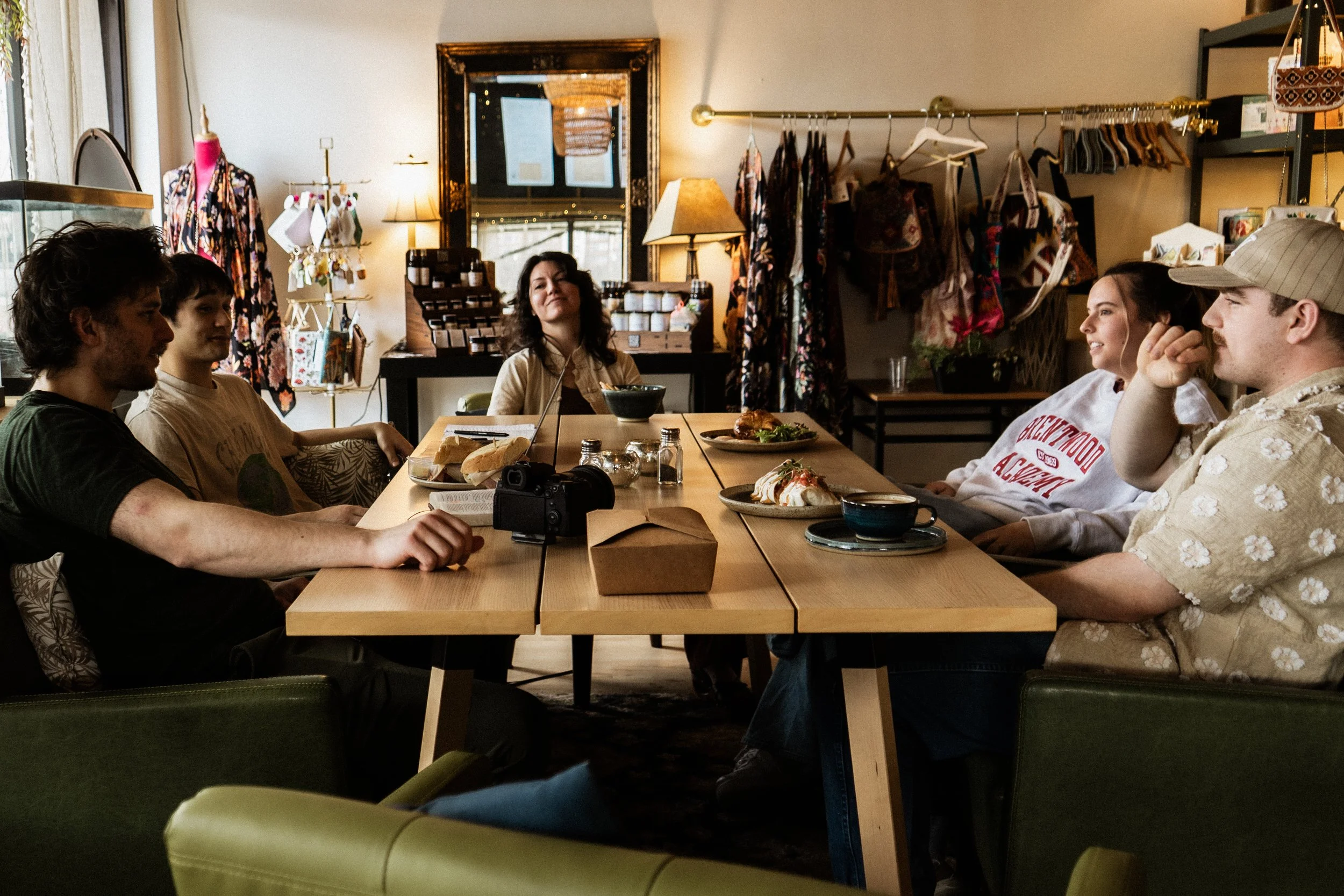 A group of five people sitting around a wooden table in a cozy café or restaurant, engaging in conversation and enjoying food. The setting includes shelves with accessories and clothing, lamps, and a mirror on the wall.