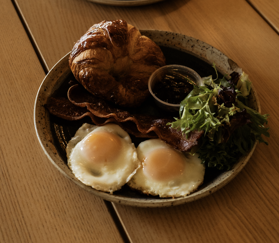 Breakfast plate with two sunny-side-up eggs, bacon, a croissant, a small bowl of syrup, and a side salad on a ceramic plate.