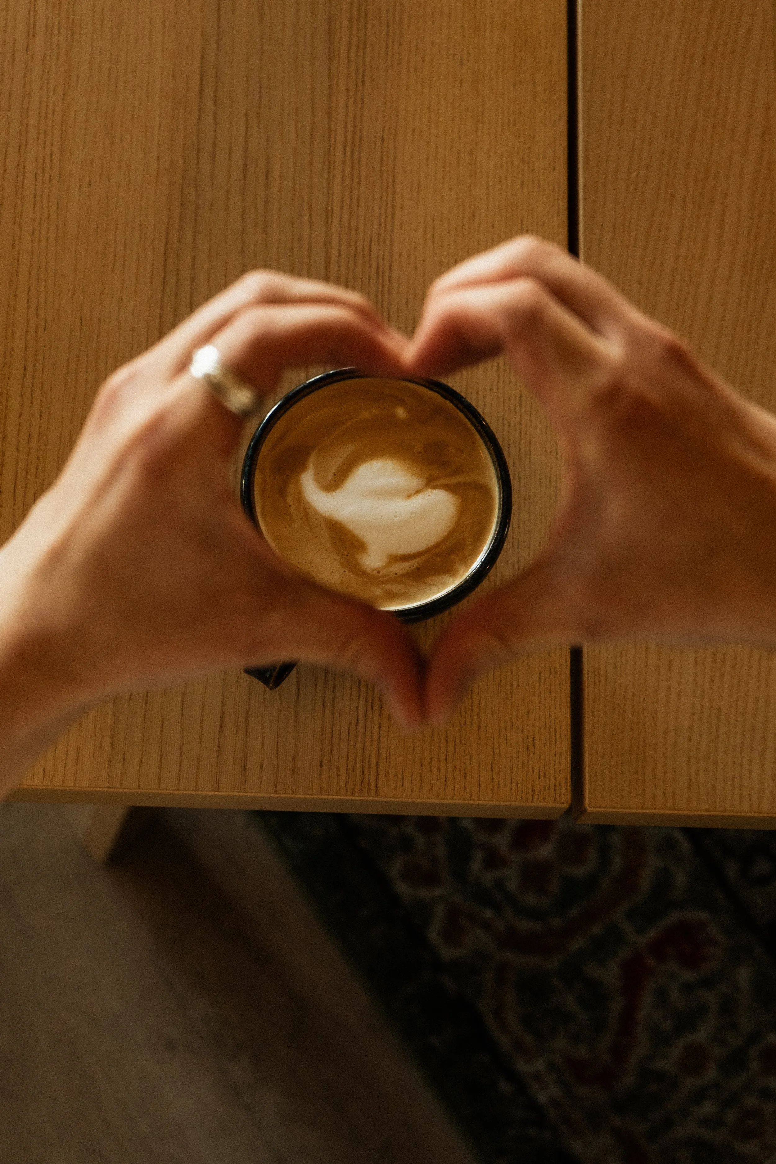 A person making a heart shape with their hands around a coffee mug on a wooden table.
