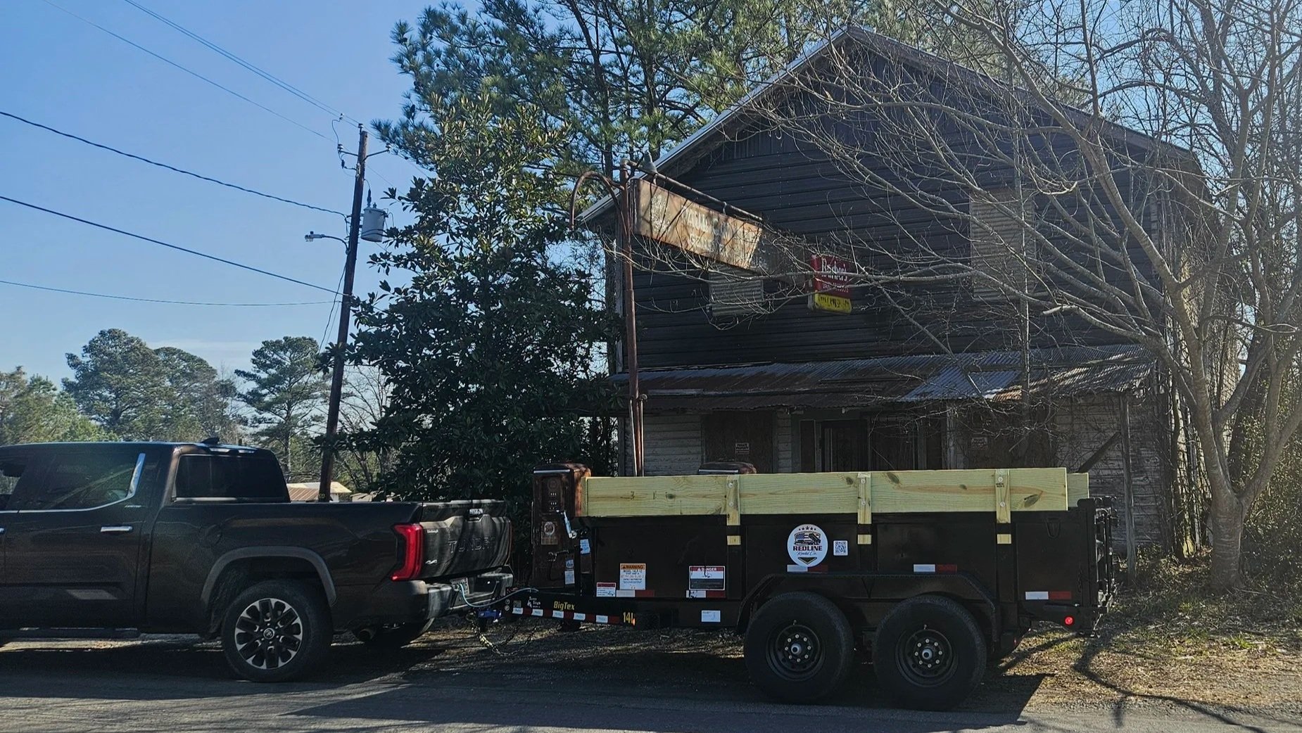 Black pickup truck attached to a trailer carrying building materials parked on a residential street with trees and an old house in the background.