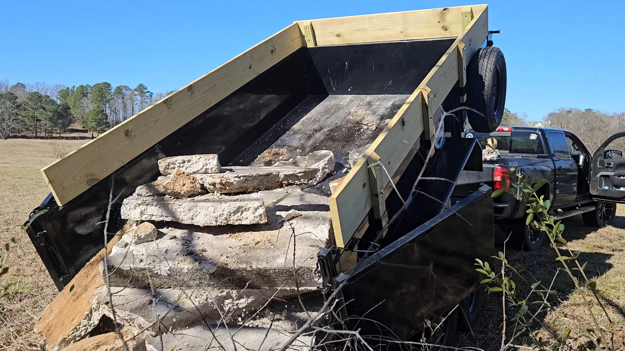 A black pickup truck with the bed tilted, dumping a load of rocks and debris onto the ground in an open field. The truck is outdoors under a clear blue sky with trees in the background.
