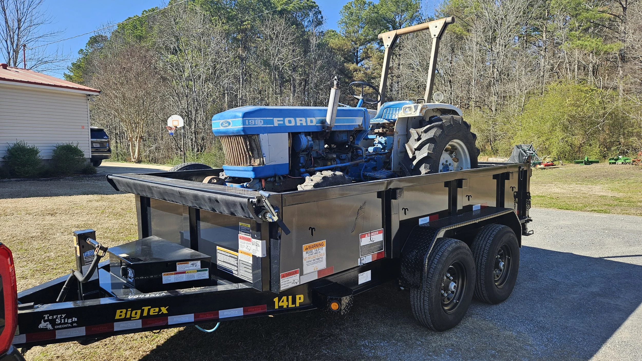 A blue vintage Ford tractor loaded on a black utility trailer in a residential yard with a house, driveway, and trees in the background.