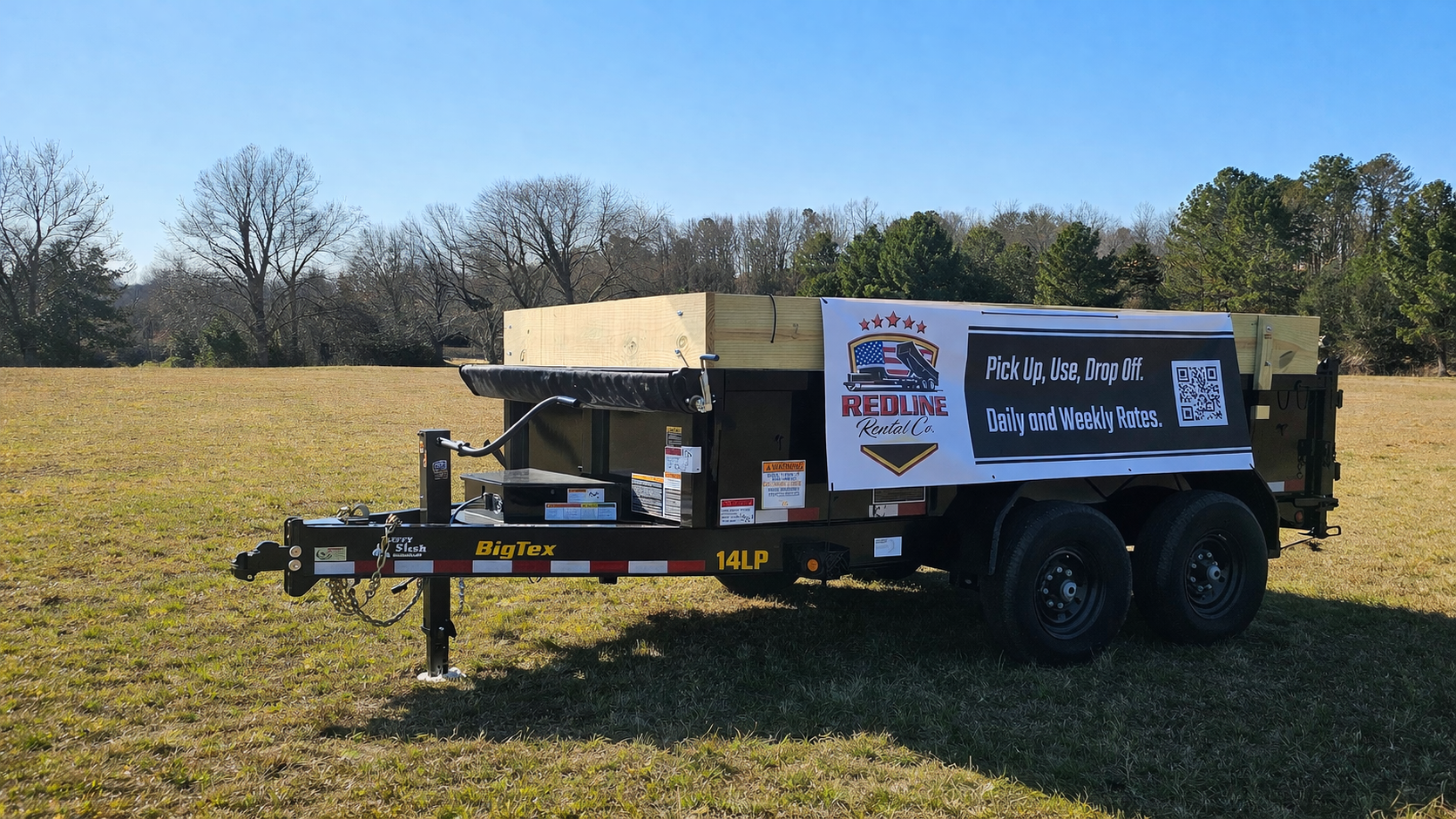 A utility trailer with a sign advertising Redline Rental Co., placed in an open grassy field with trees in the background and a clear blue sky.