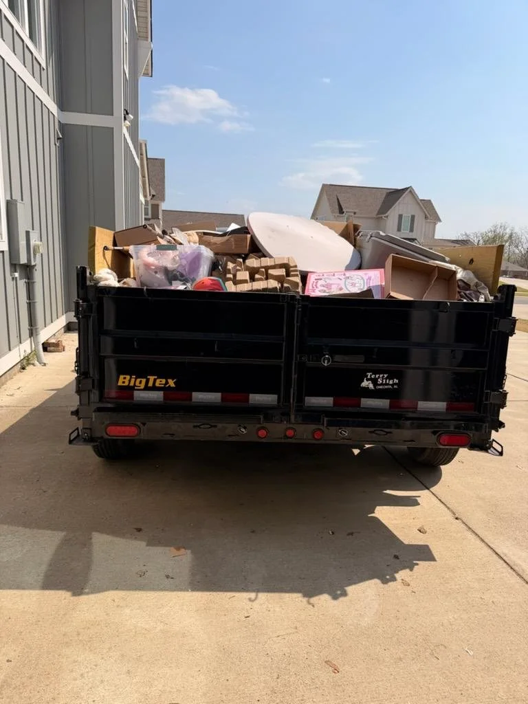 Trailer filled with assorted trash and discarded household items parked on a driveway, with residential buildings and a clear sky in the background.