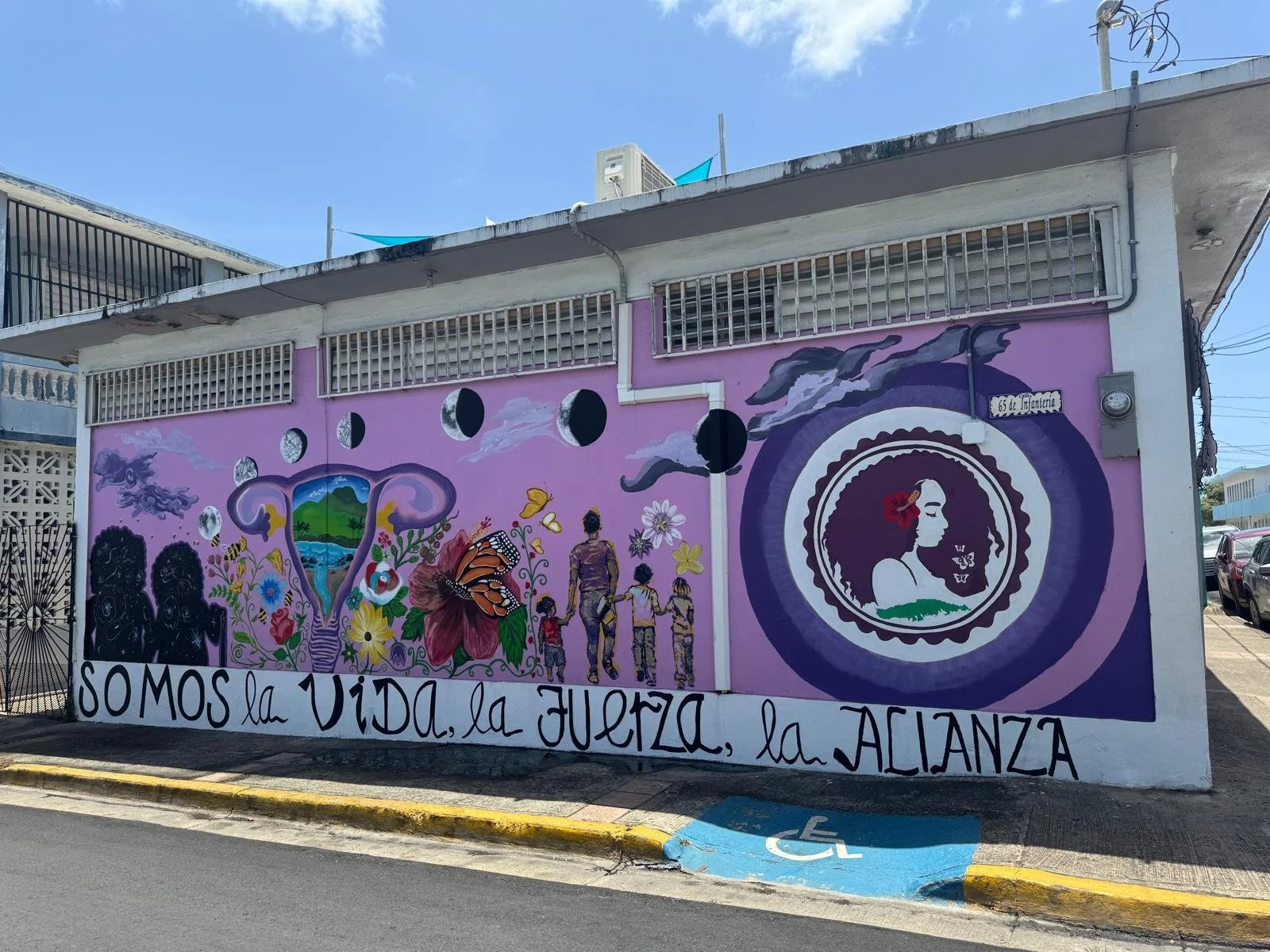 Colorful painted pink mural with flowers, butterflies, a family holding hands, and a woman's face in profile with a flower in her hair, on a building wall. The text reads 'Somos la vida, la fuerza, la alianza'.
