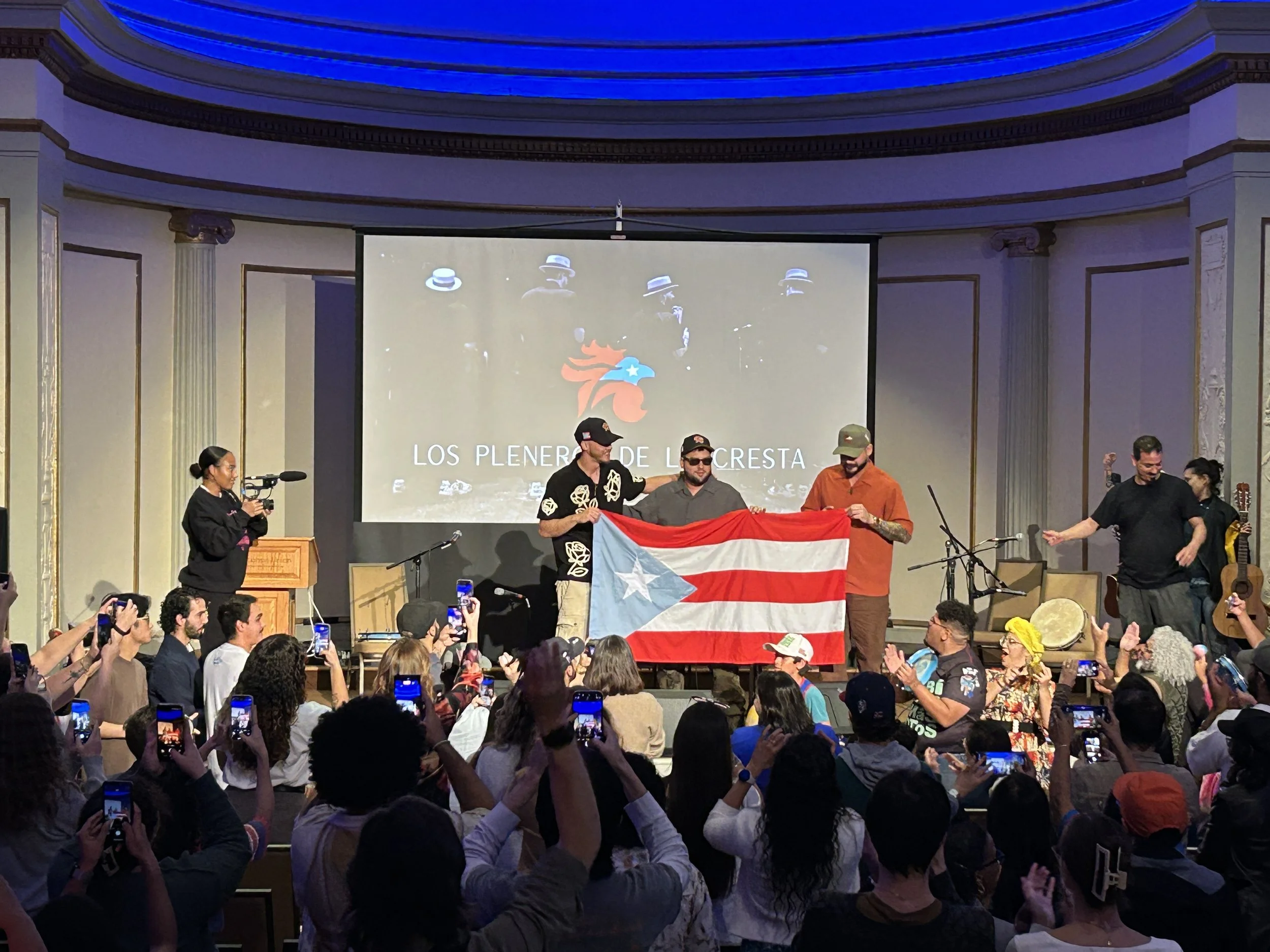People on stage holding a Puerto Rican flag during an event with a large audience taking photos, a woman with a camera, and a projection on the screen behind them showing a logo and the text 'Los Pleneros de la Cresta'.