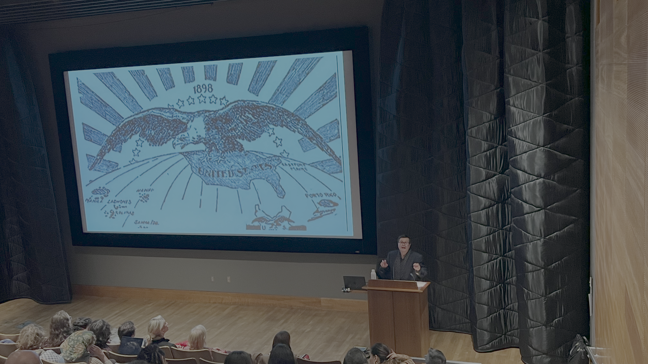 A woman presenting in a lecture hall with a large projection of a vintage map of the United States on the screen behind her.
