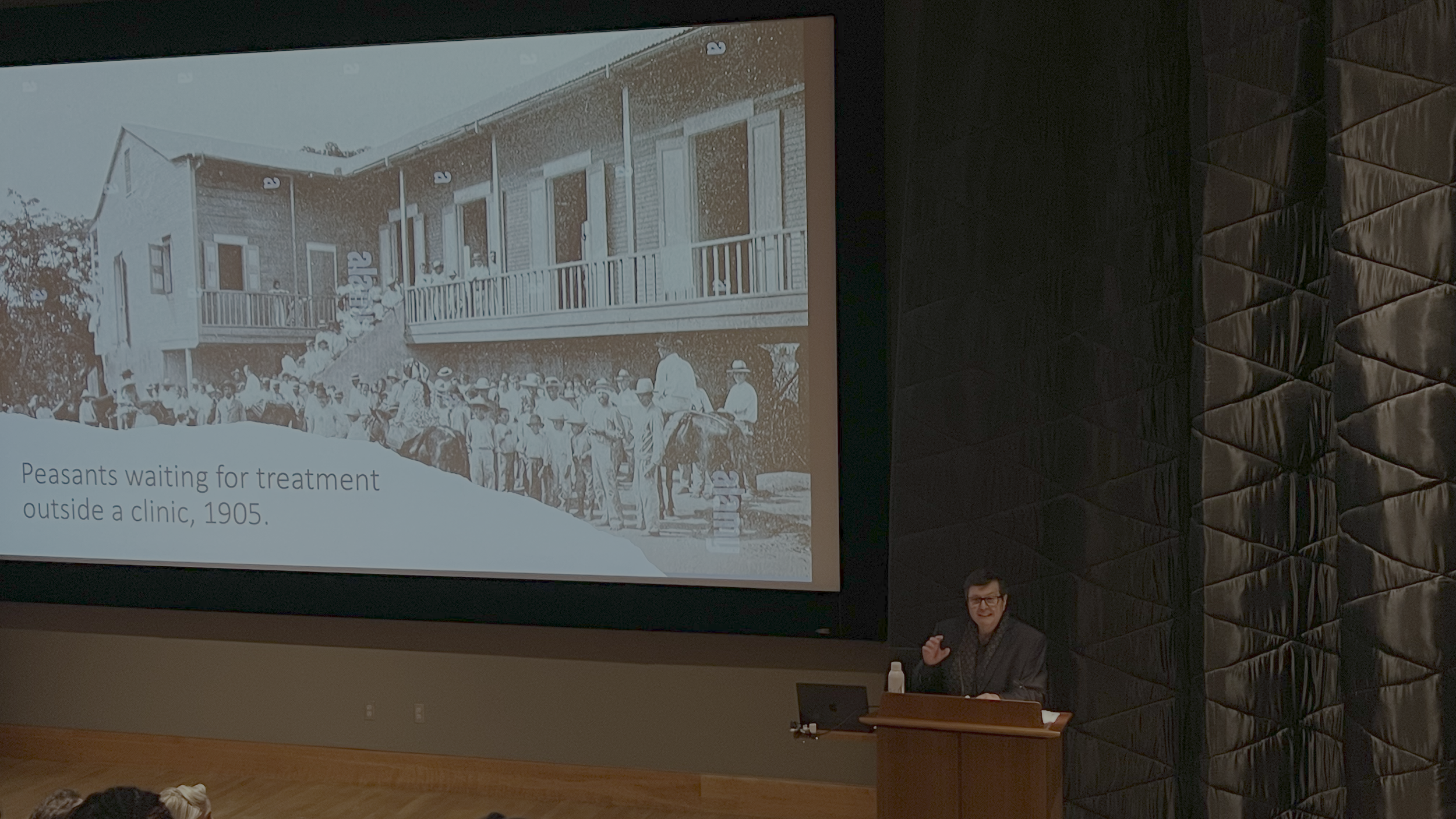 Francisco Scarano presenting a slide with a vintage photo of jíbaro Puerto Rican patients waiting outside a doctor's office.