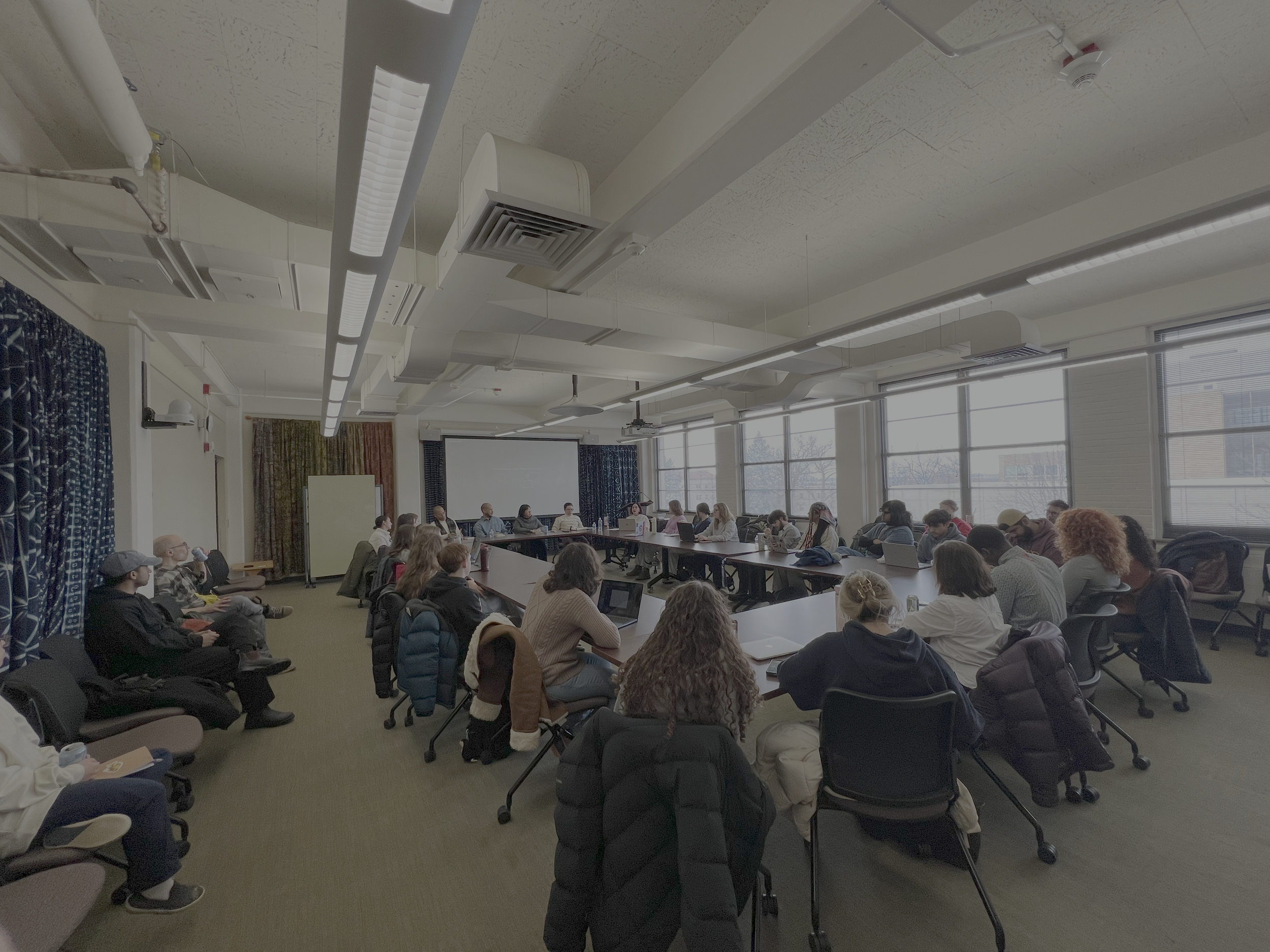 A large conference room filled with people seated around a U-shaped table, attending a presentation or meeting, with windows along one wall and ceiling lights.