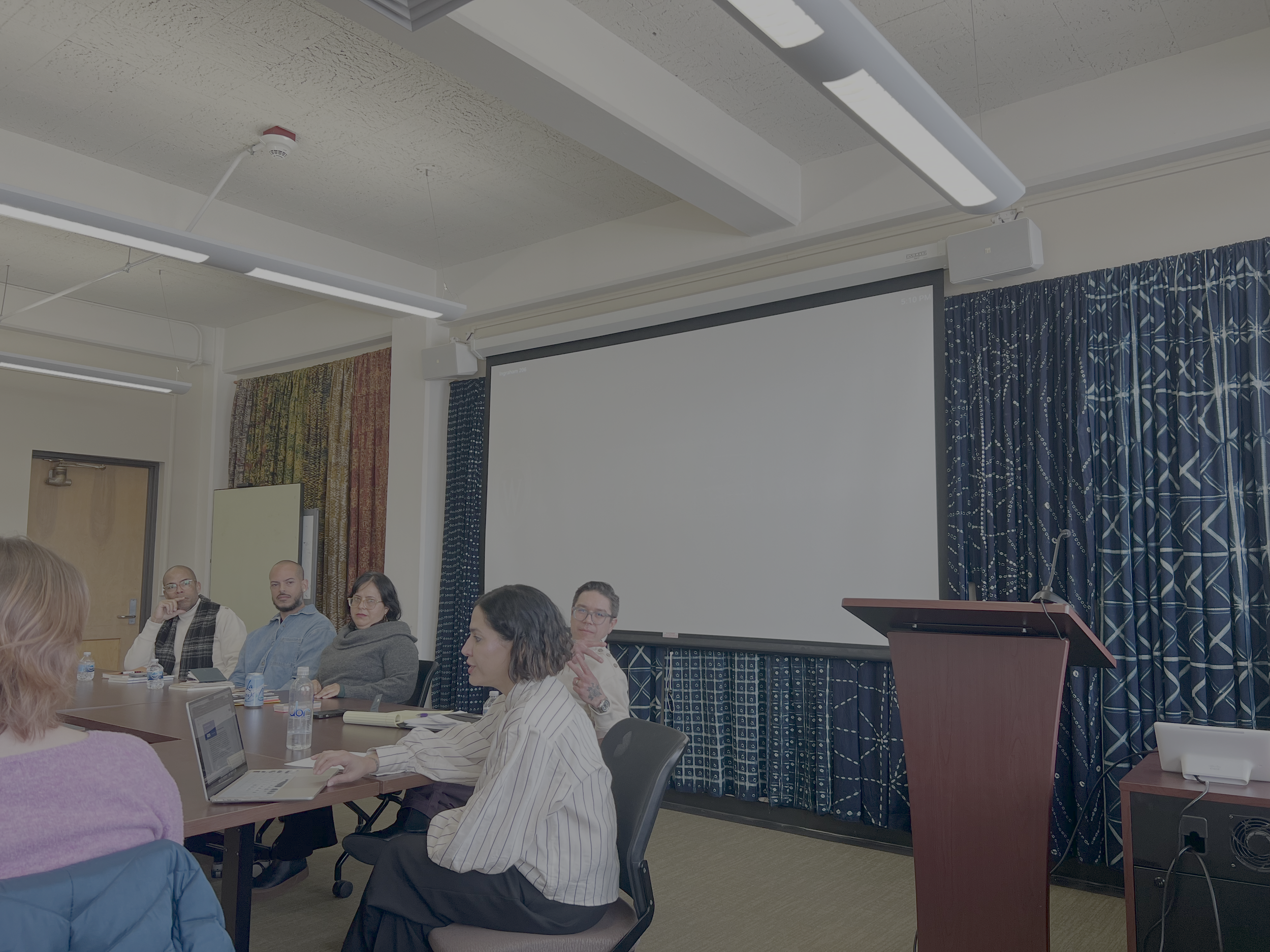 A group of diverse people sitting around a conference table in a meeting room, some taking notes or typing on laptops, with a large projection screen and curtains in the background.