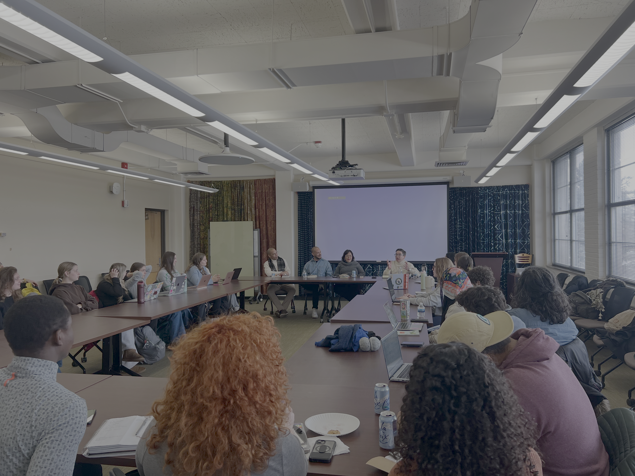 A large group of people sitting around a long conference table in a meeting room, with a presentation screen and a presenter at the front.