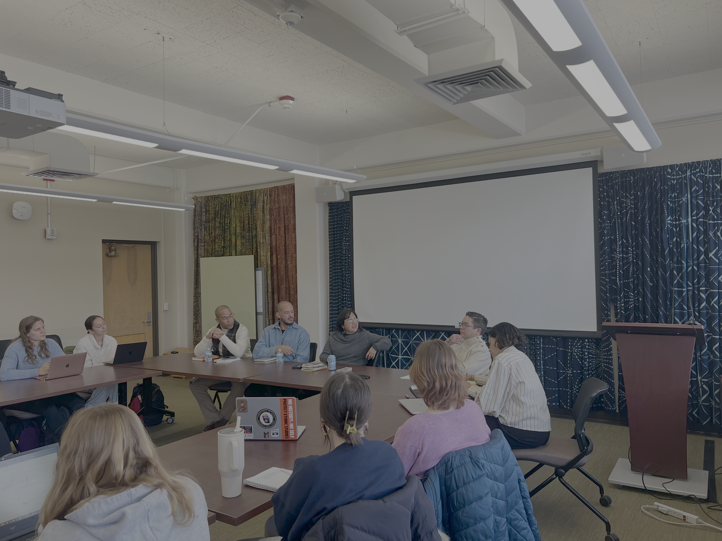 People attending a meeting or class in a conference room with a projector, a large screen, and curtains.
