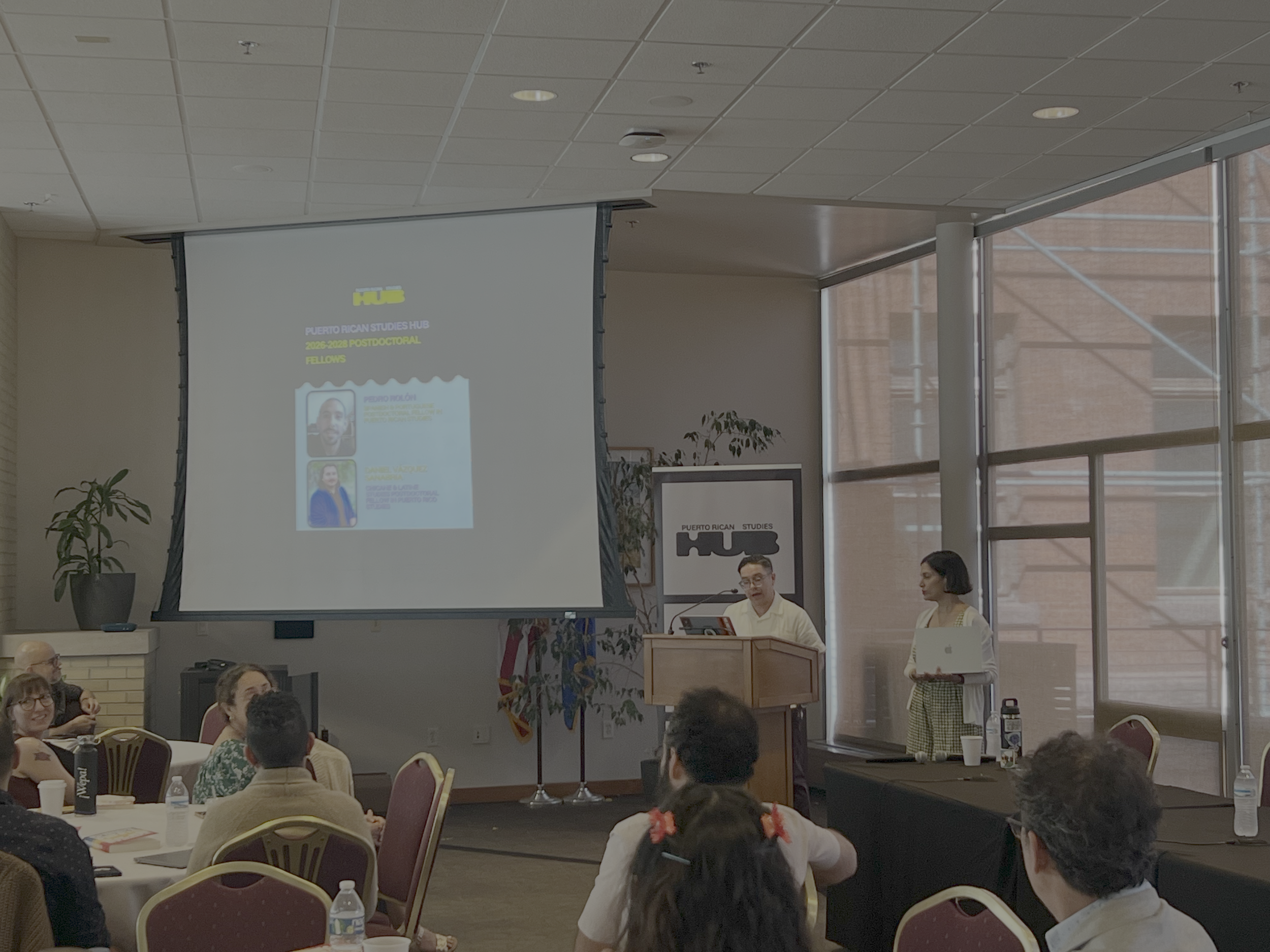 A conference room with a projector screen displaying a presentation for the Puerto Rican Studies Hub, featuring photos of speakers Pedro Muñiz and Daniel Azuela. Several attendees are seated at round tables, and a man stands near the stage speaking. 