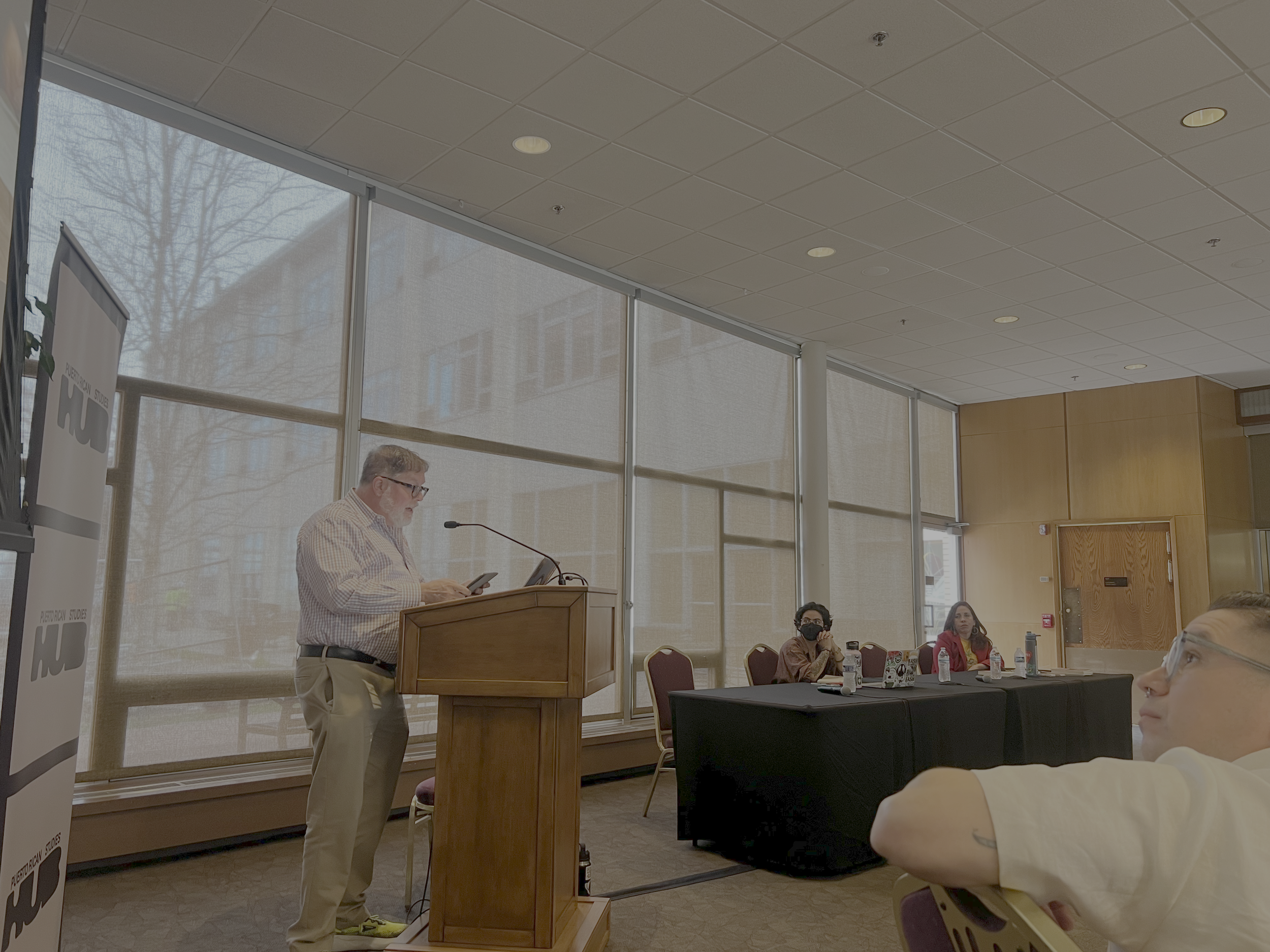 A man in a plaid shirt is standing behind a wooden podium, speaking into a microphone and looking at his phone. Several people are seated in a conference room with large windows, some wearing masks, listening to the speaker.