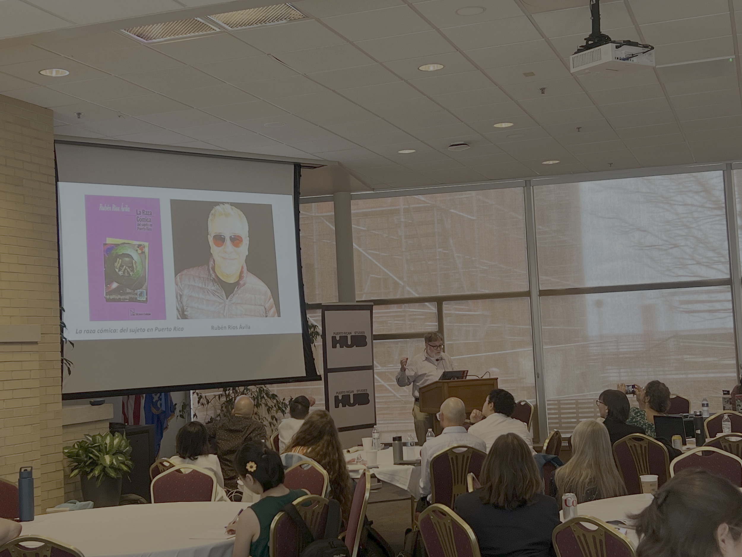 A man is giving a presentation at a seminar with many attendees. The presentation slide features an image of a person with white hair wearing sunglasses and a person in black glasses. The room has large windows showing an outdoor scene, and a project
