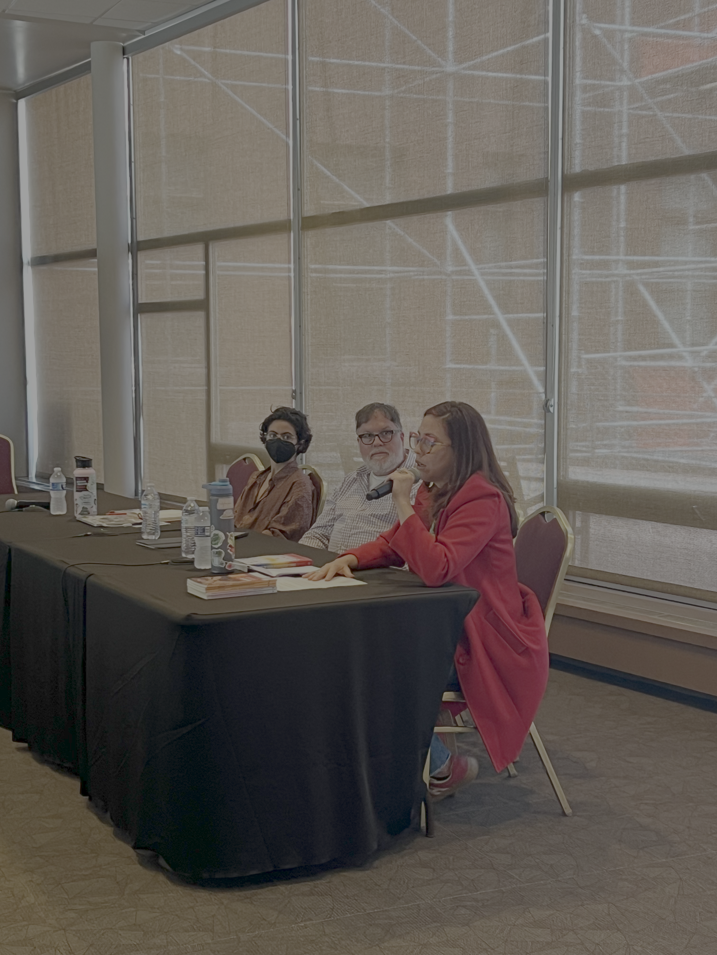 People sitting at a conference table during a panel discussion, with three women in the foreground, one woman is speaking into a microphone while others observe. The table has water bottles, notebooks, and papers, and the background shows beige windo
