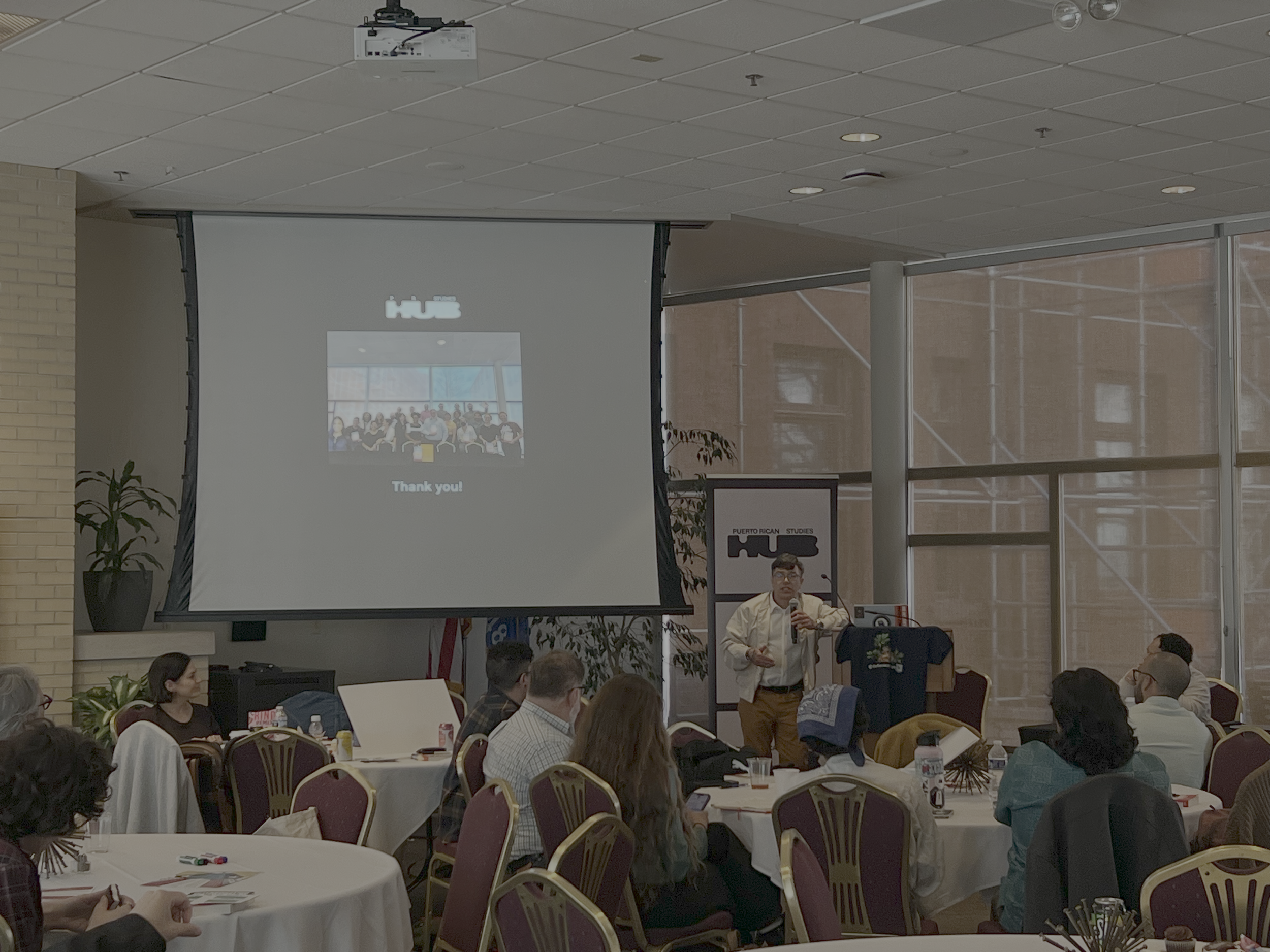 A conference room with a standing speaker, a group of people seated at round tables, a large screen displaying a group photo with the words 'Thank you!', and a windowed wall in the background.