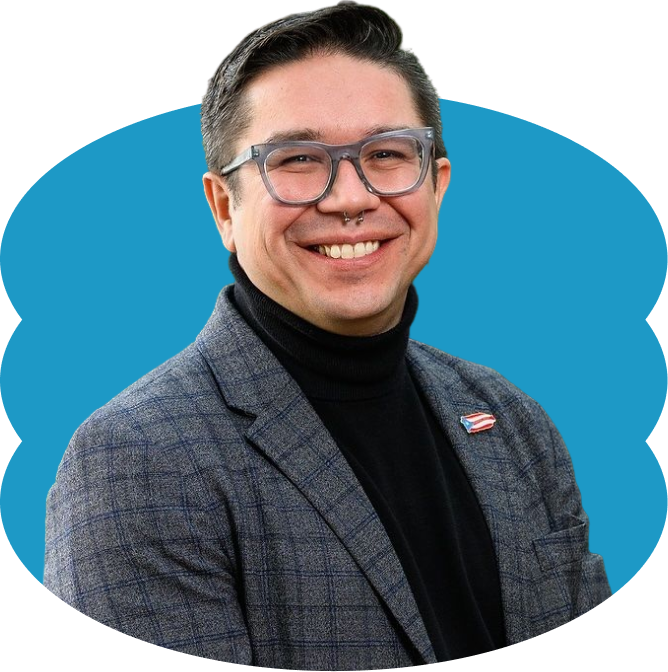 Jorell Meléndez-Badillo, a smiling man with short hair, glasses, and a nose ring, wearing a black turtleneck and grey suit jacket with a Puerto Rican flag pin, placed over a scalloped blue oval shape.