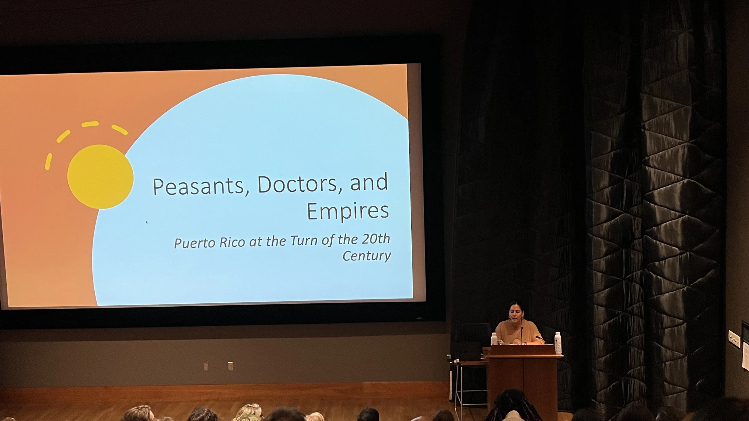 A woman is presenting a lecture in an auditorium. The presentation slide on the large screen behind her is titled 'Peasants, Doctors, and Empires' with a subtitle 'Puerto Rico at the Turn of the 20th Century'.
