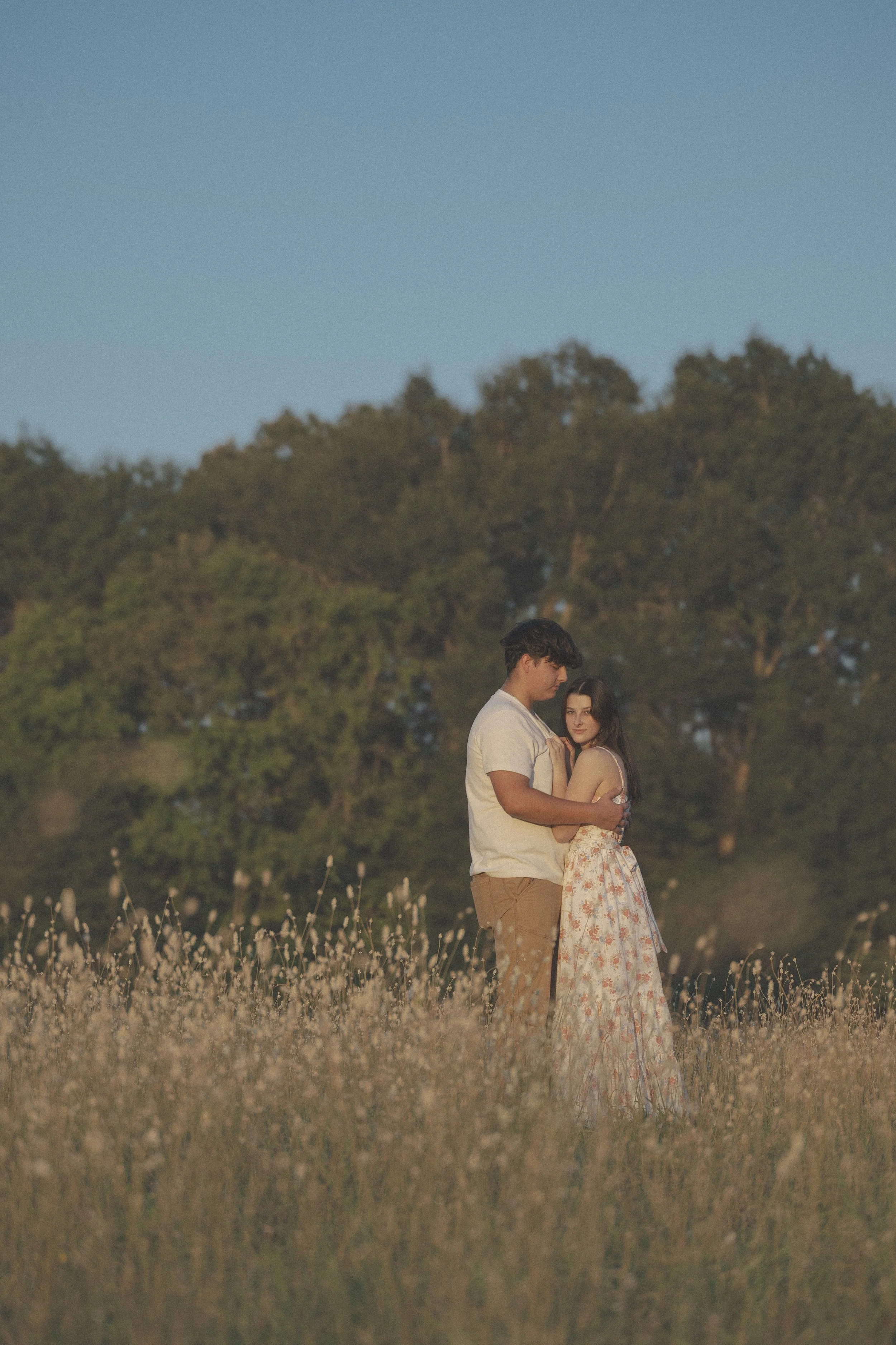 A young couple standing close together in a field of tall grass, with trees and a clear sky in the background.