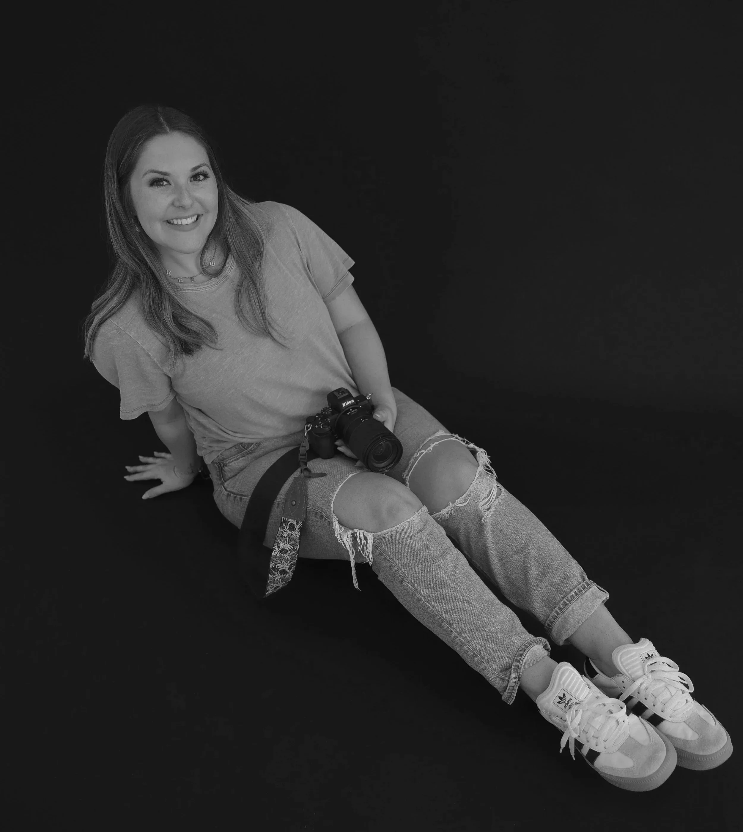 Black and white photo of a young woman sitting on the floor with a camera in her lap, smiling at the camera, dressed casually in a t-shirt and ripped jeans, wearing white sneakers, with a dark plain background.