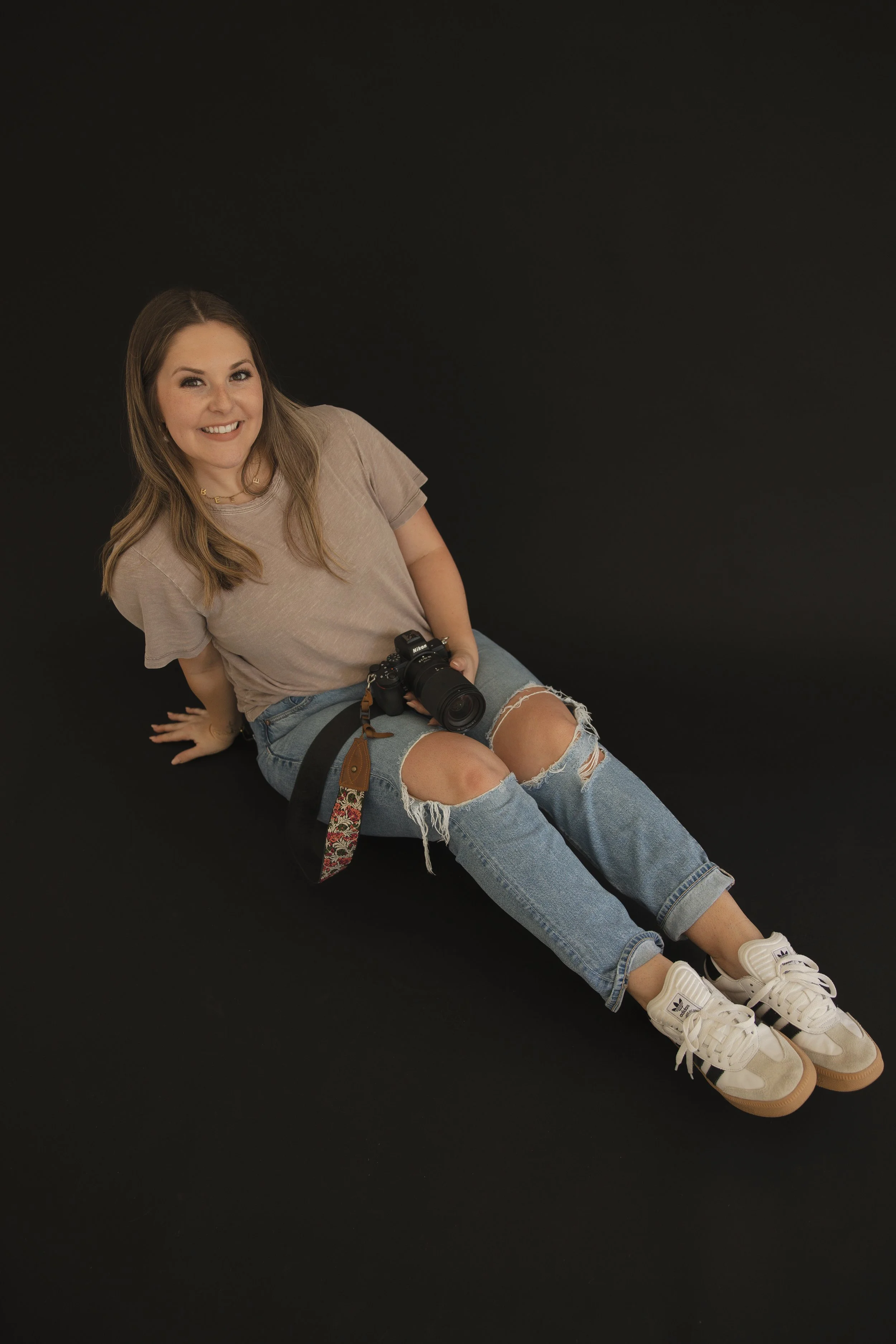 Young woman sitting on a black surface against a black background, smiling, with a camera on her lap, wearing a beige t-shirt, ripped jeans, and Adidas sneakers.