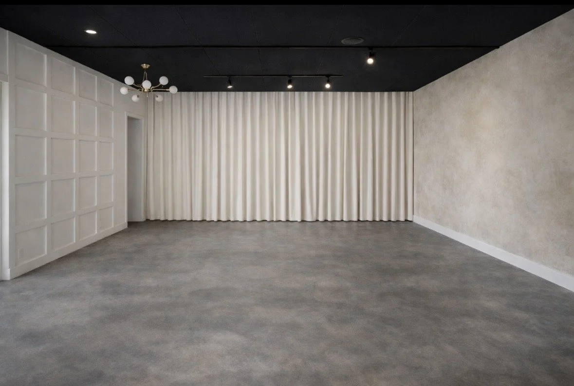 Empty room with gray carpet, white paneled wall on the left, white textured wall on the right, white curtains covering the back wall, black ceiling with small sports lights and a modern chandelier.