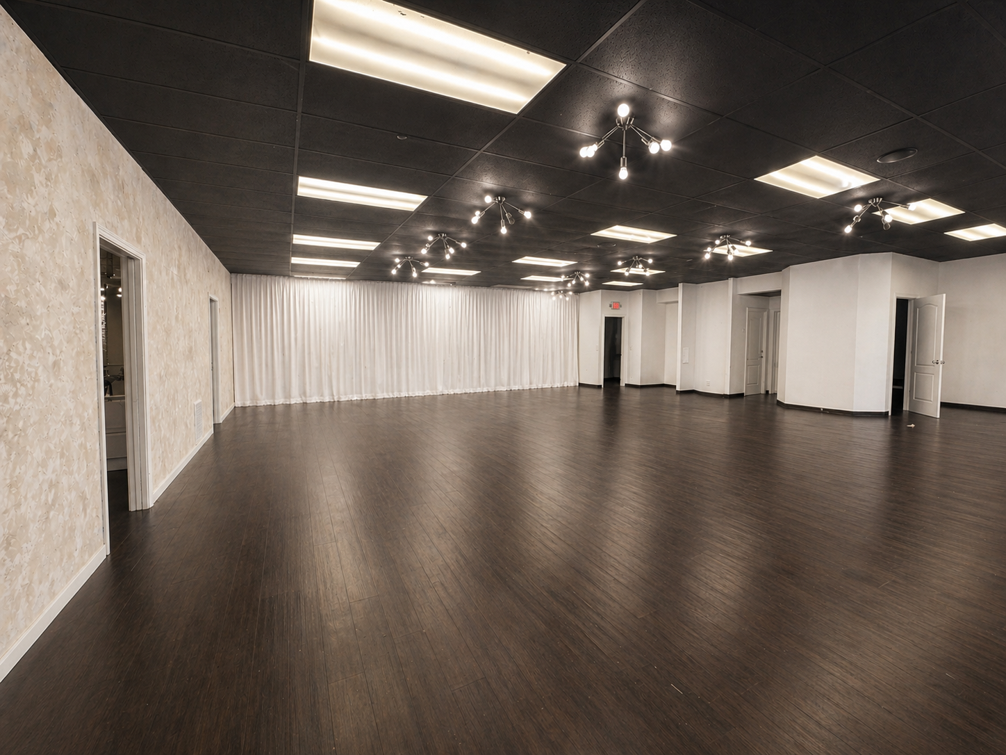 Empty event space or dance hall with dark wood flooring, white walls, a curtain on one wall, and multi-bulb chandeliers on a black ceiling with rectangular lights.
