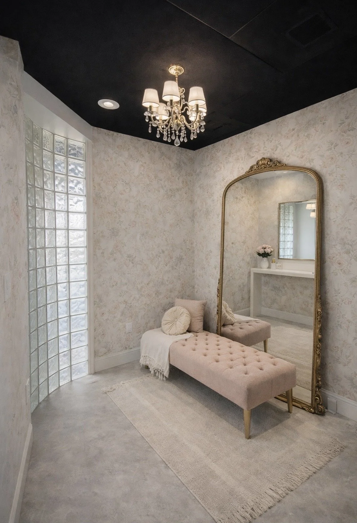 Elegant room with floral wallpaper, a large ornate mirror, a pink tufted bench, a white throw blanket, a round pillow, a chandelier, and a glass block wall letting in natural light.