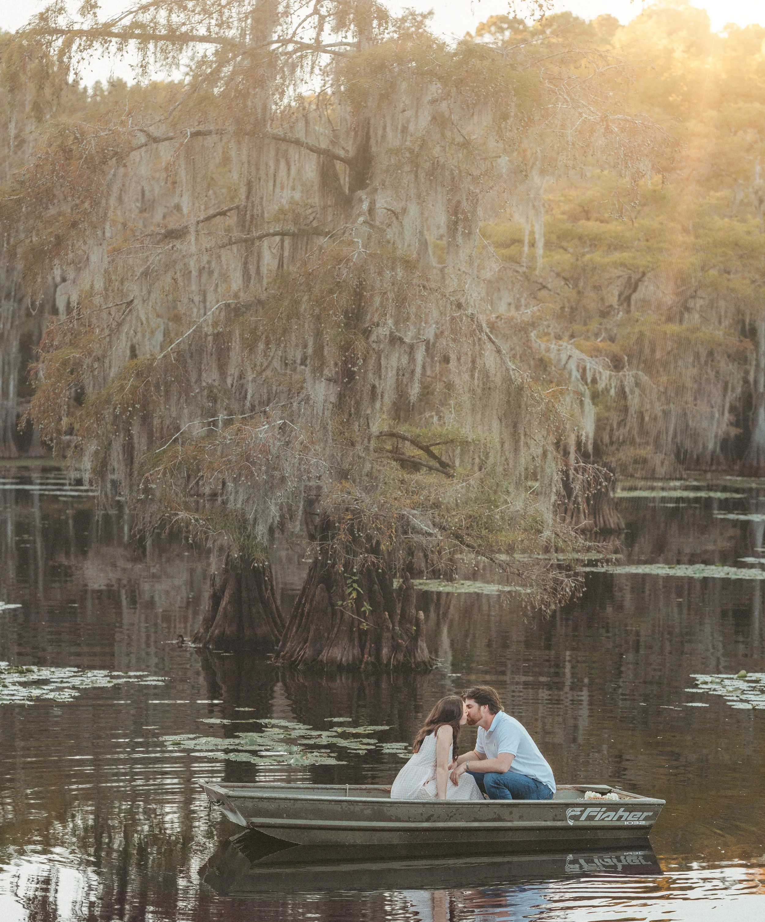 A couple sits closely in a boat on a calm lake, sharing a kiss, with a large moss-covered tree and autumn trees in the background.