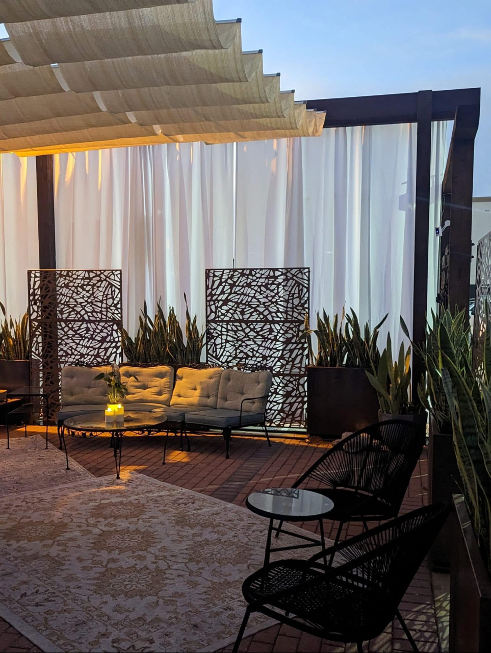 Outdoor seating area with a cushioned sofa, black wire chairs, small glass tables, potted plants, patterned screen dividers, and overhead fabric shading, illuminated by warm lighting at dusk.
