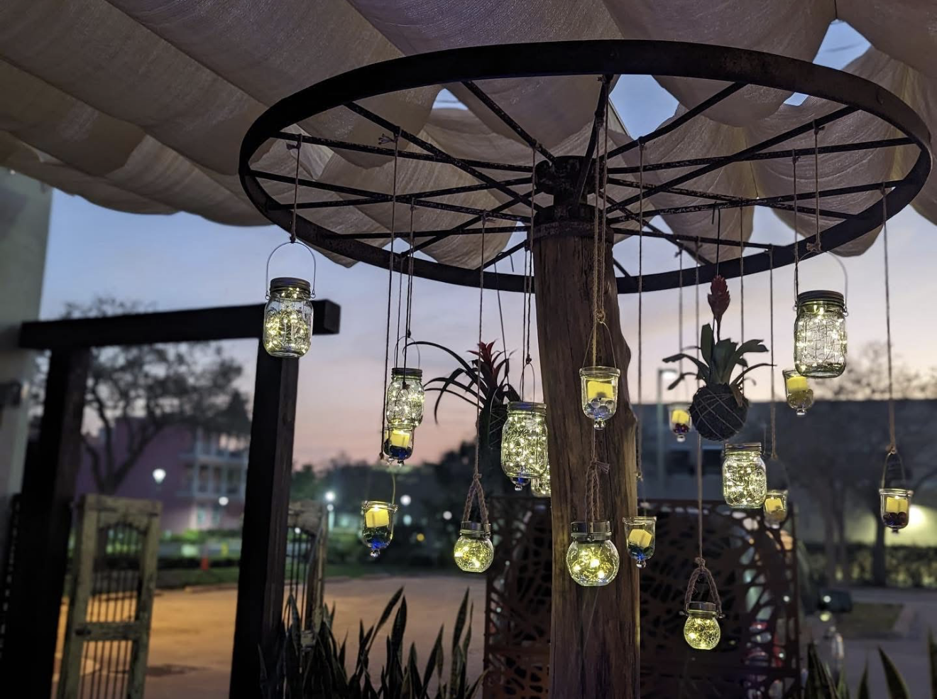 Outdoor decoration with hanging jars filled with string lights and small flowers, hanging from a wooden structure at dusk.