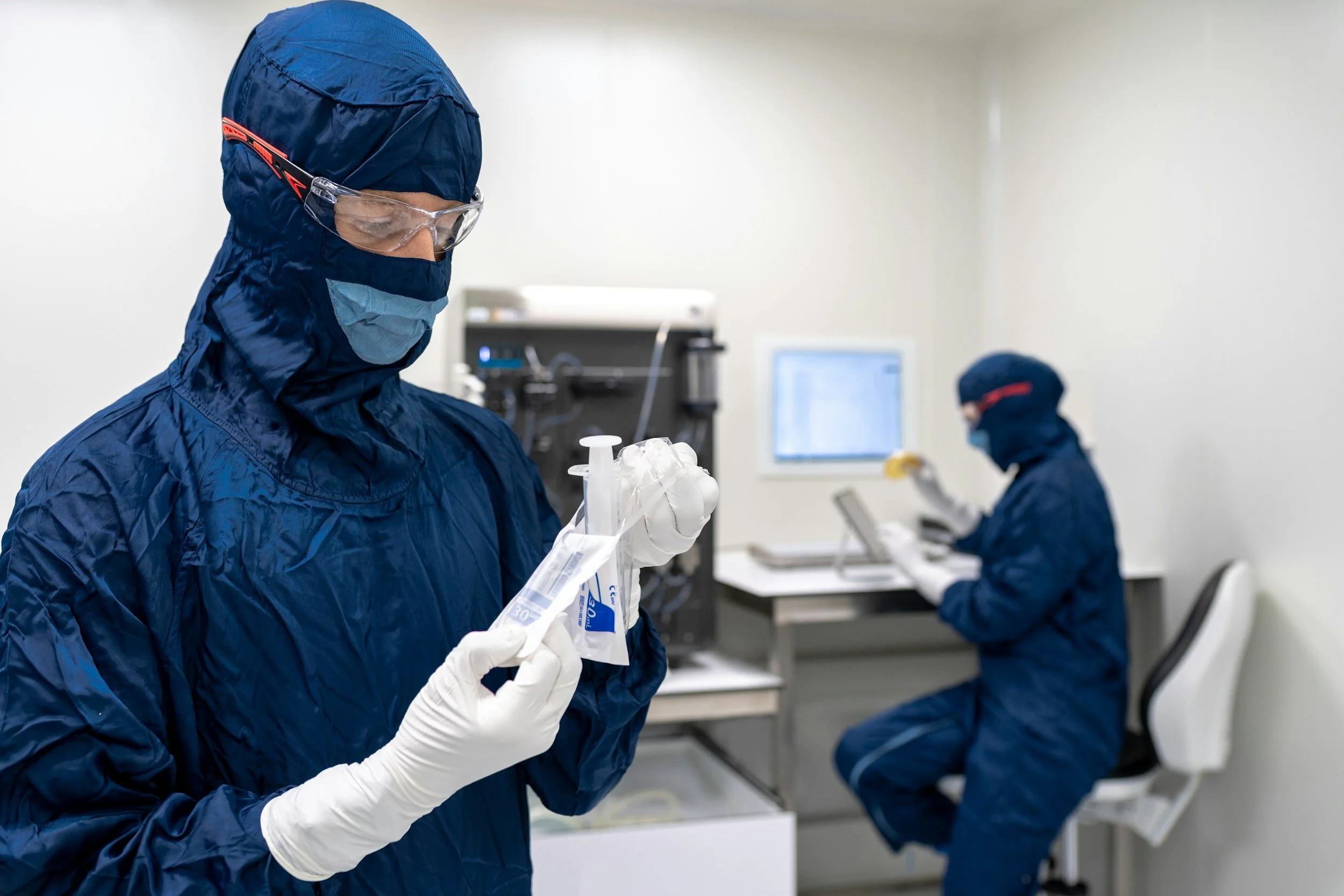 Two scientists in protective blue suits, gloves, masks, and goggles working in a laboratory. One scientist is holding a sample in a bag, while the other is seated at a desk working on a computer.