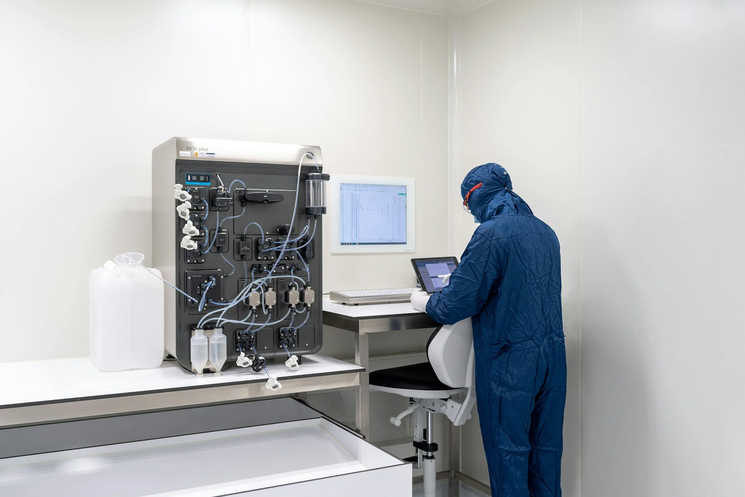 A scientist in full protective gear operating a medical device and a tablet in a sterile laboratory.