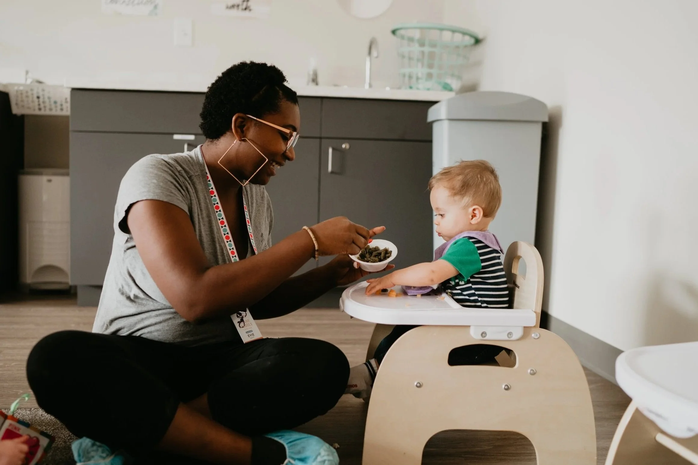 A woman with glasses sitting on the floor feeding a young child in a high chair in a kitchen.