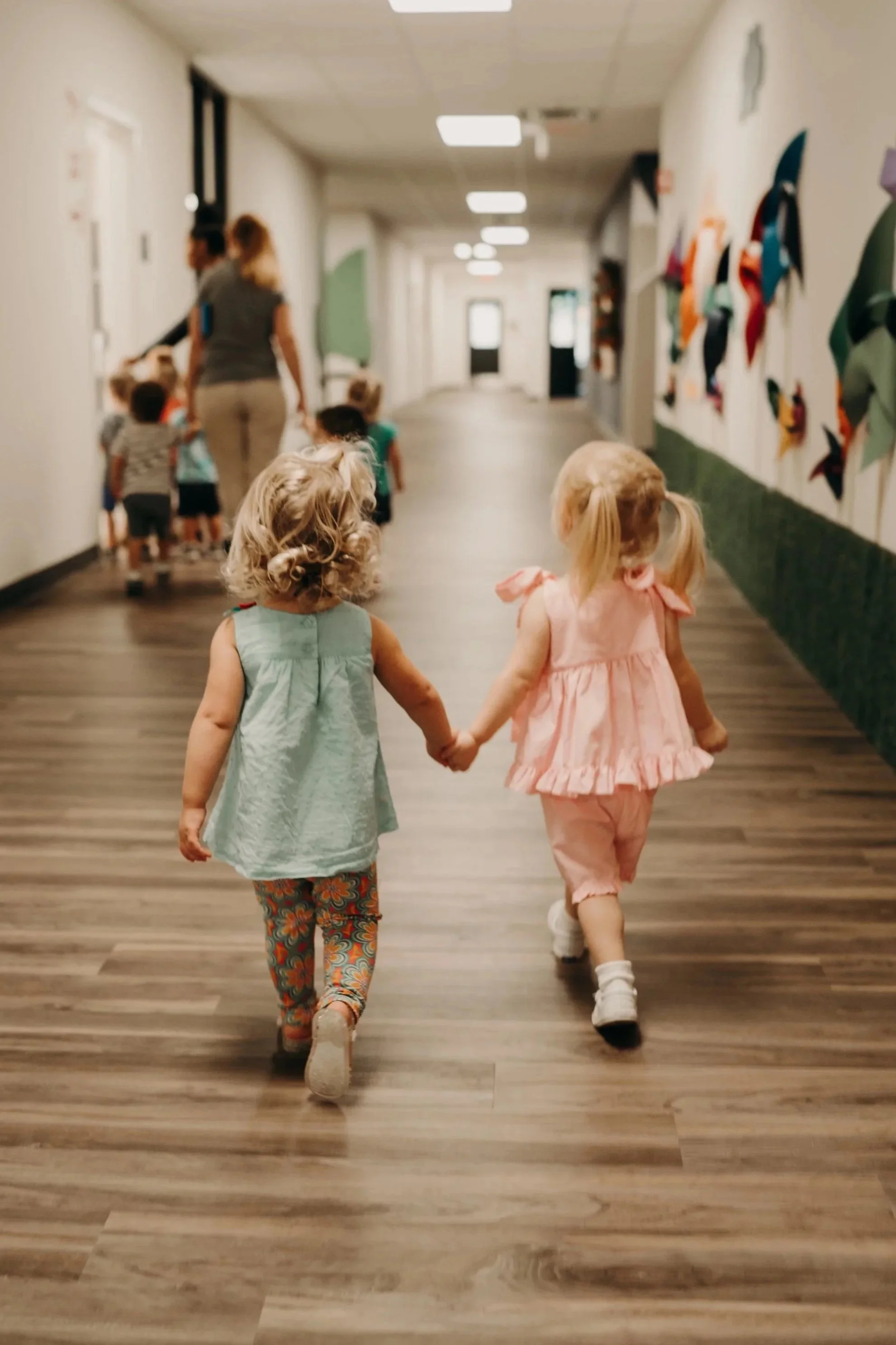 Two young girls holding hands walk down a hallway at a preschool or daycare, with other children and caregivers in the background.