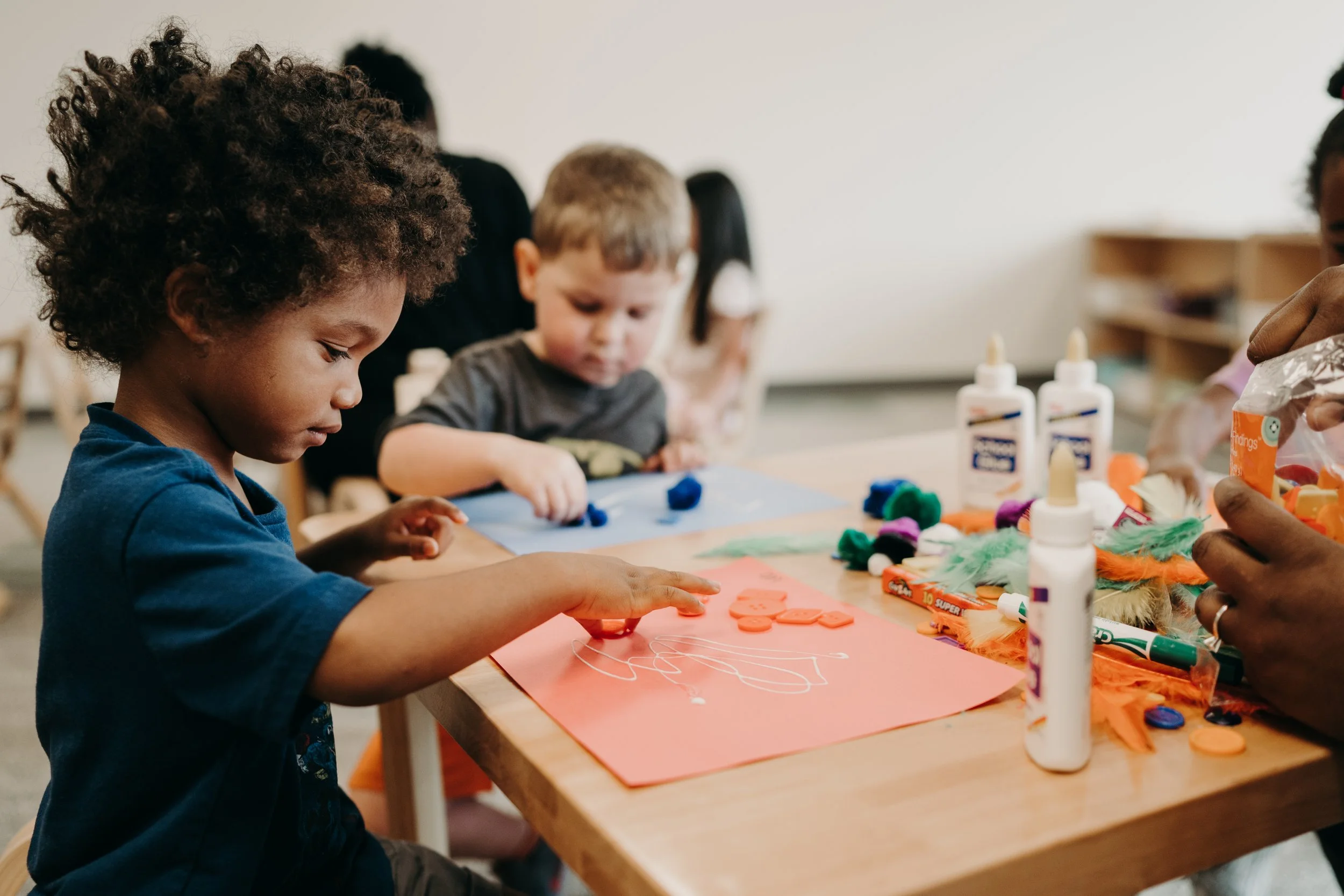 Children engaging in an arts and crafts activity at a table with glue, colorful foam shapes, yarn, and paper.
