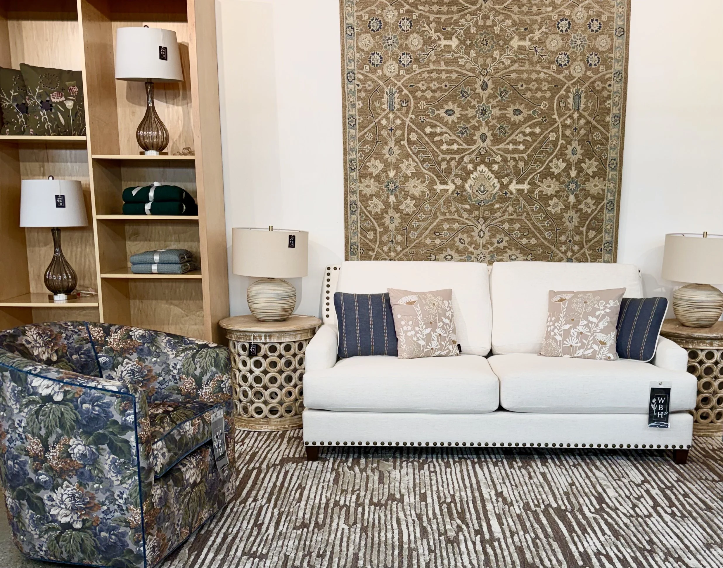 Living room display with a white sofa, a floral armchair, a patterned rug, a large decorative wall hanging, and a wooden bookshelf with lamps, folded textiles, and decorative items.