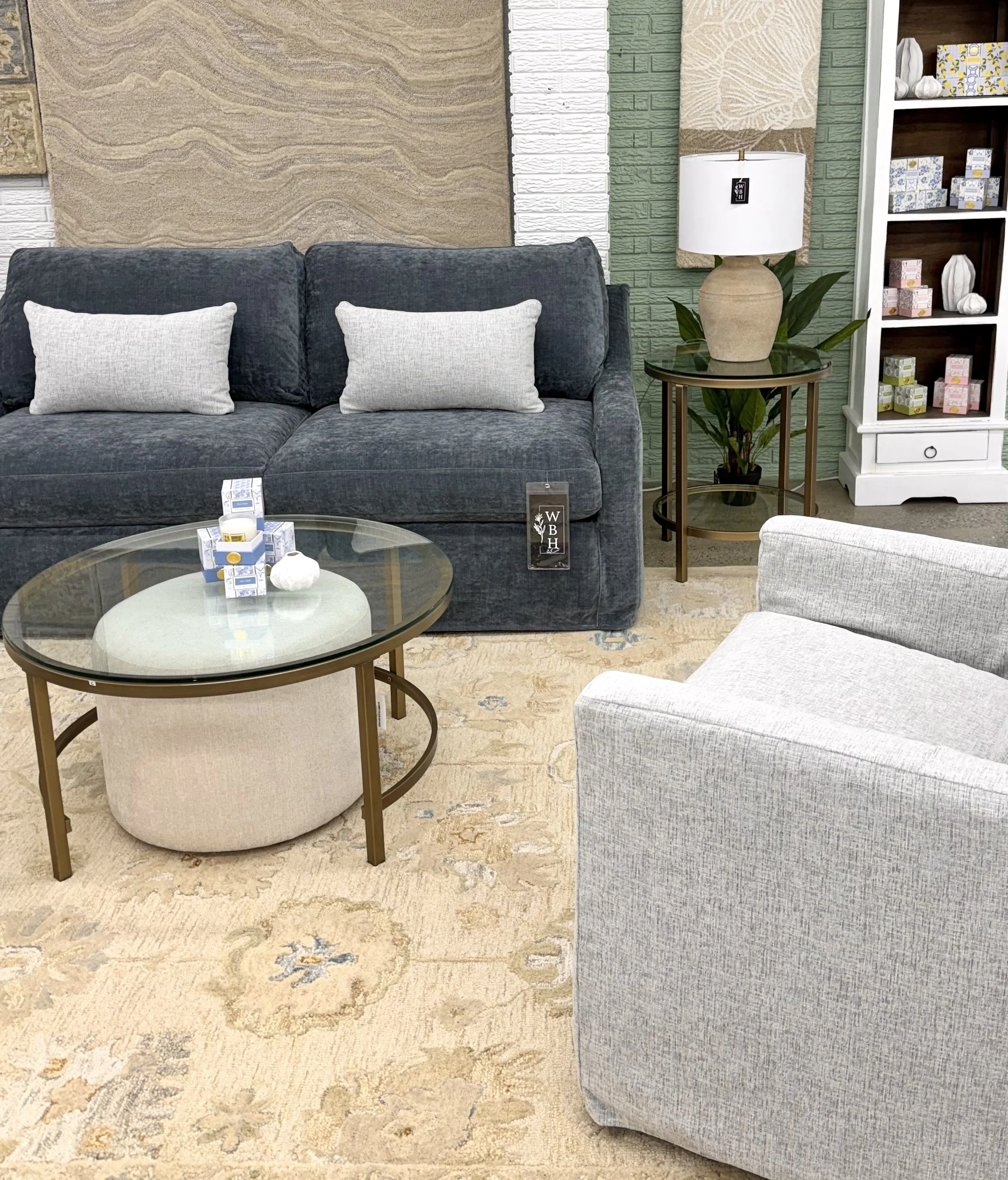 A living room with a navy blue sofa decorated with two white pillows, a glass-top coffee table with items on it, a side table with a table lamp and a plant, a white bookshelf with decor items, and a beige and green textured wall with artwork.