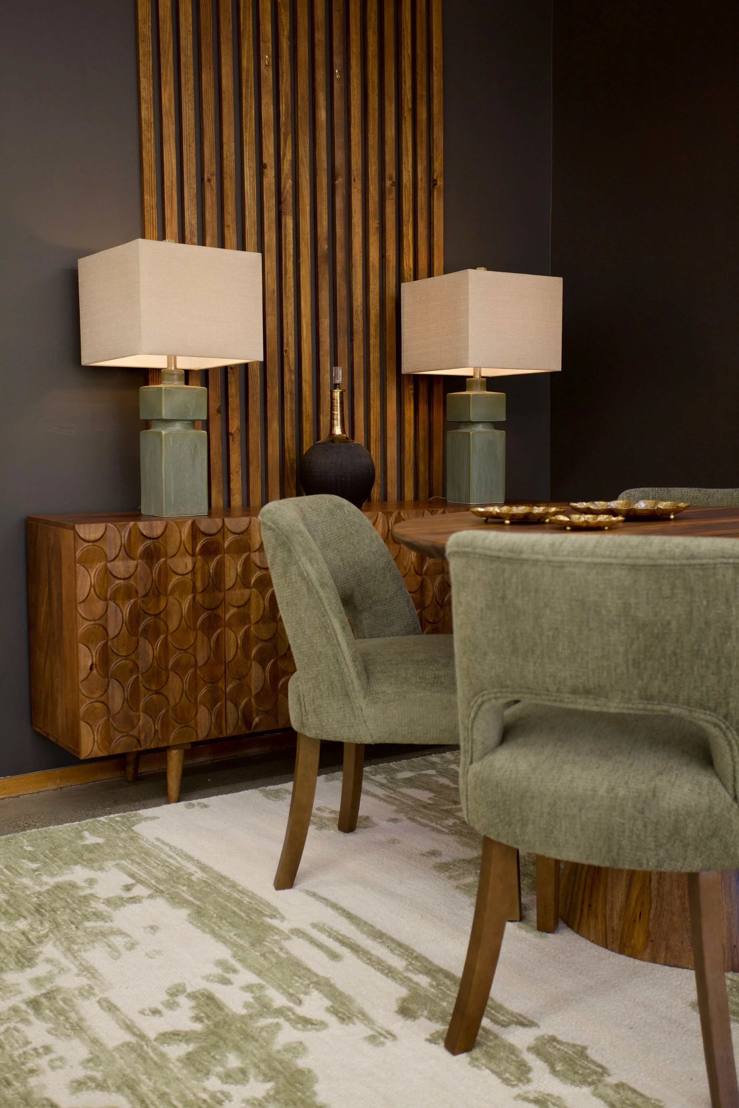 Interior of a room with decorative furniture, including a wooden sideboard with circular pattern, two matching lamps with beige shades on the sideboard, one black and gold decorative vase, a light green armchair with wooden legs, a wooden dining tabl