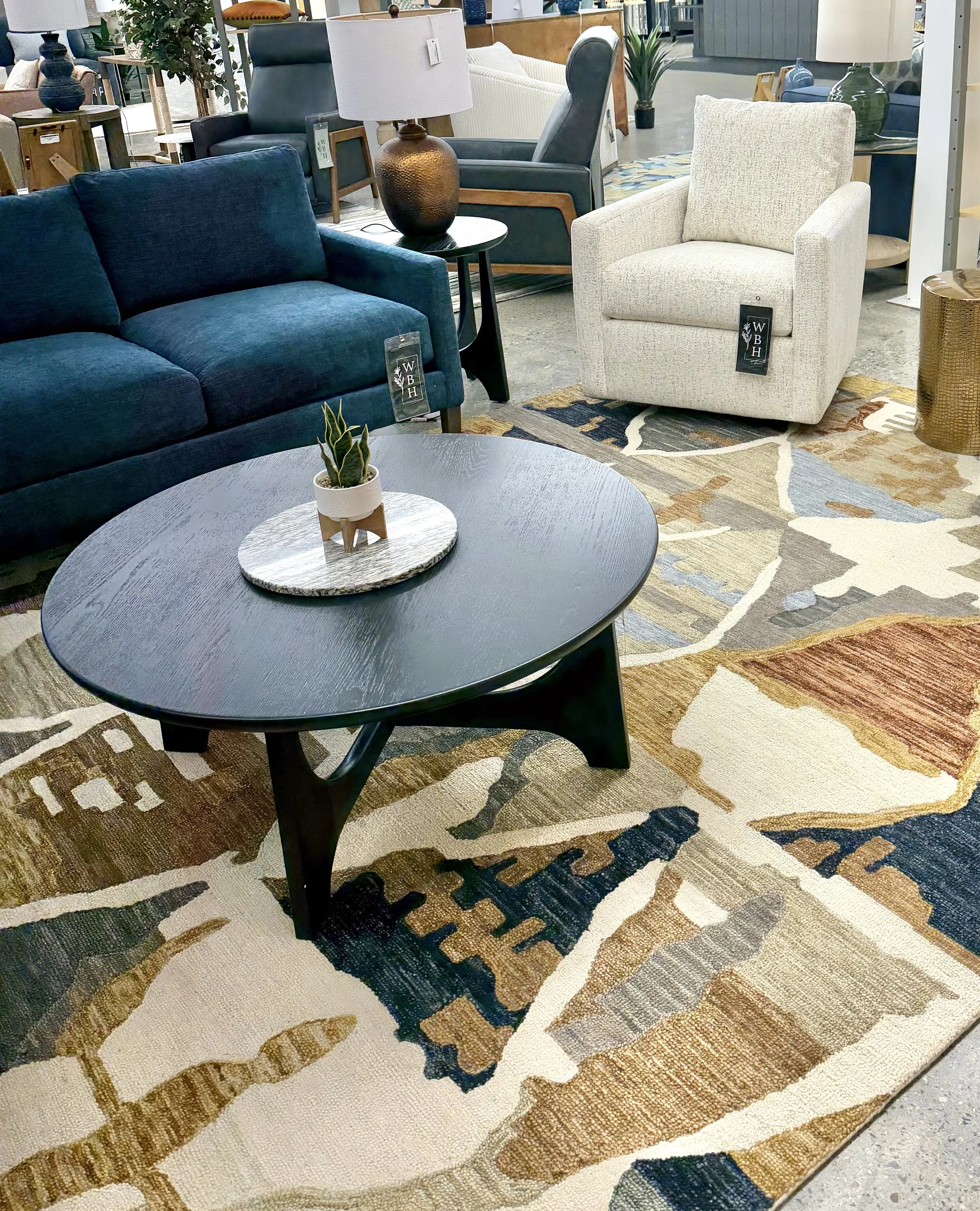 A furniture display in a store with a blue sofa, a white armchair, a black side table with a small plant, and various lamps and decorative items, all arranged on a colorful patterned rug.