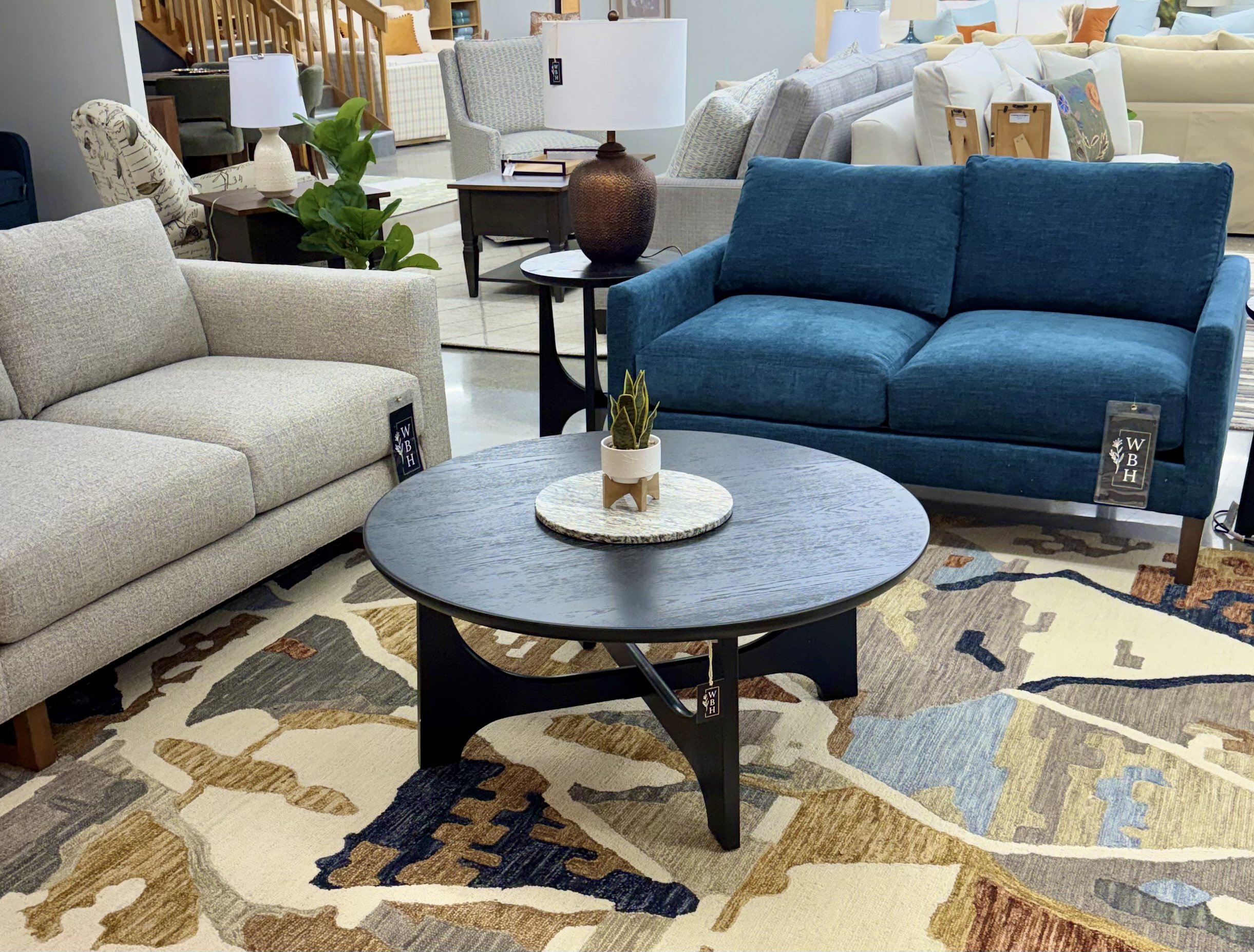 A showroom display of various sofas and furniture pieces arranged on a multicolored mosaic rug. In the foreground, a round black coffee table with a small potted plant is visible. Behind, a beige sofa and a bright blue sofa are arranged with other furniture items and lamps in the background.